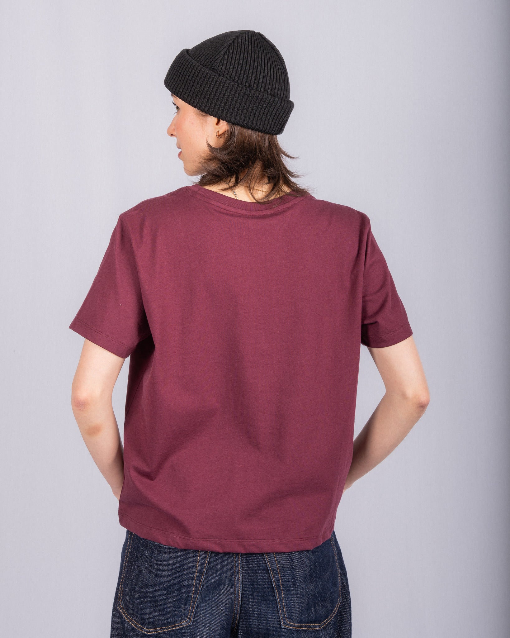 Person in a black beanie and maroon t-shirt facing away, standing against a light gray background.