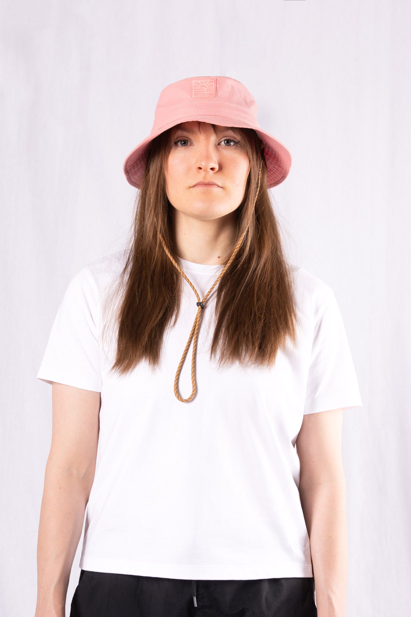 A person wearing a pink bucket hat, white t-shirt, and black pants stands against a plain white background.