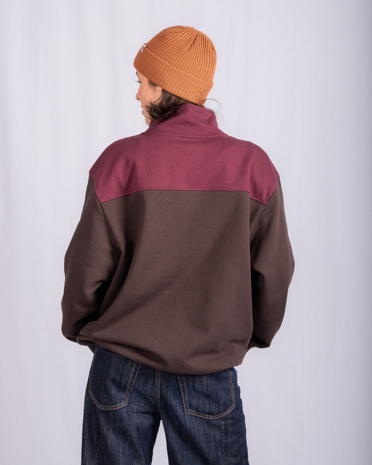 Person wearing a brown and maroon jacket, orange beanie, and jeans, facing away from the camera.
