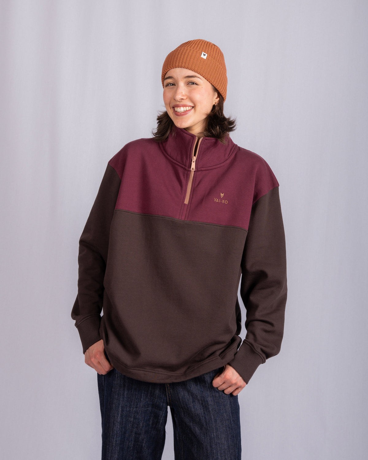 A smiling person in a brown beanie and two-tone pullover stands with hands in pockets against a gray background.