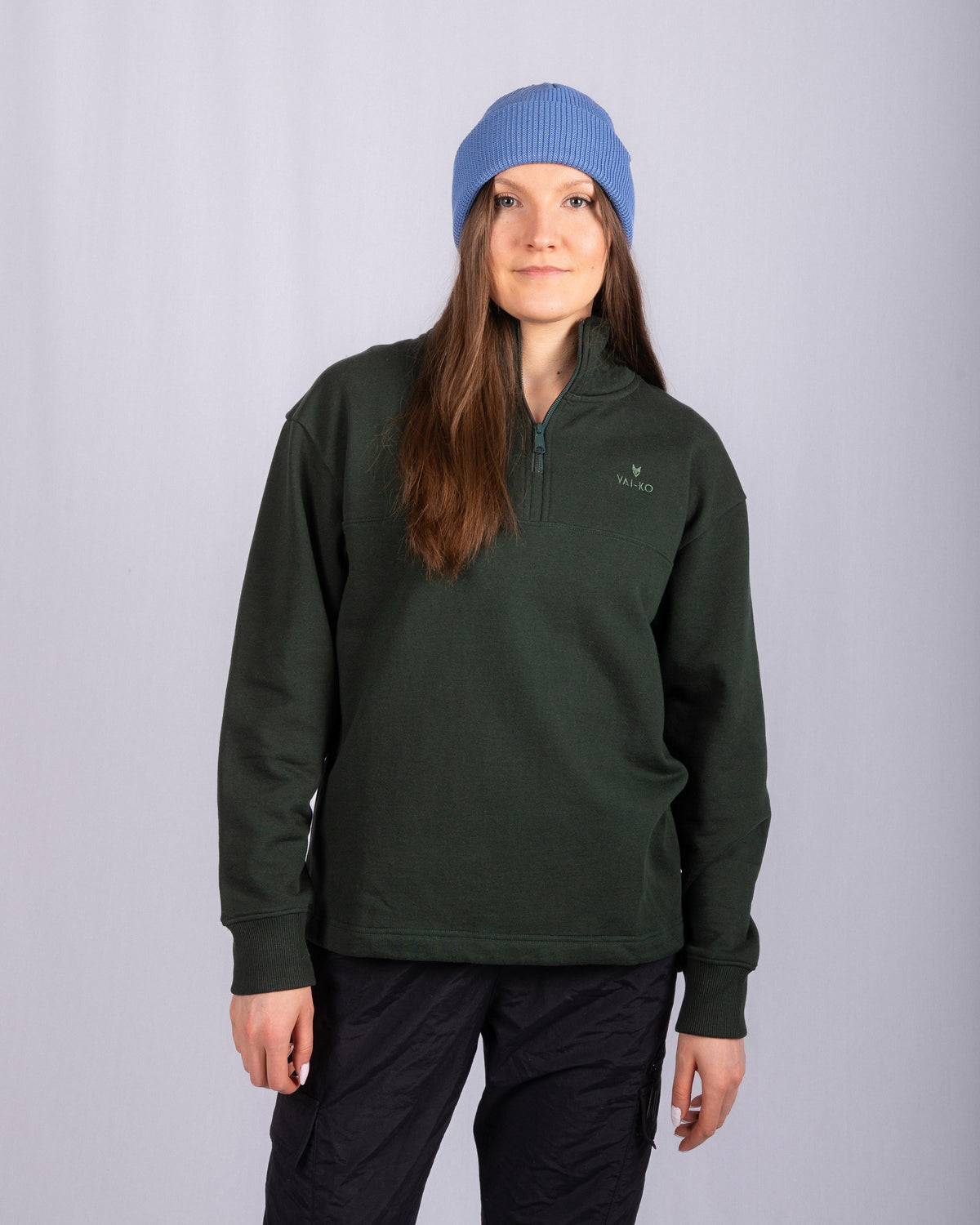 Woman in a blue beanie, dark green pullover, and black pants stands against a plain light background.