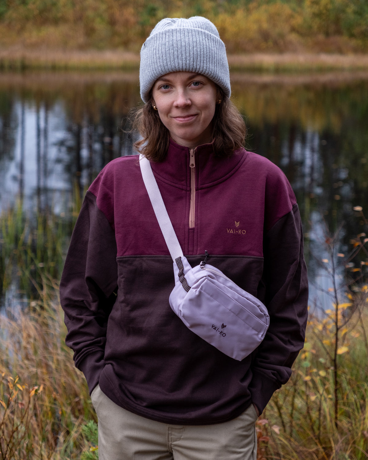 A person wearing a gray beanie, a maroon and brown quarter-zip sweater, and a light purple crossbody bag.