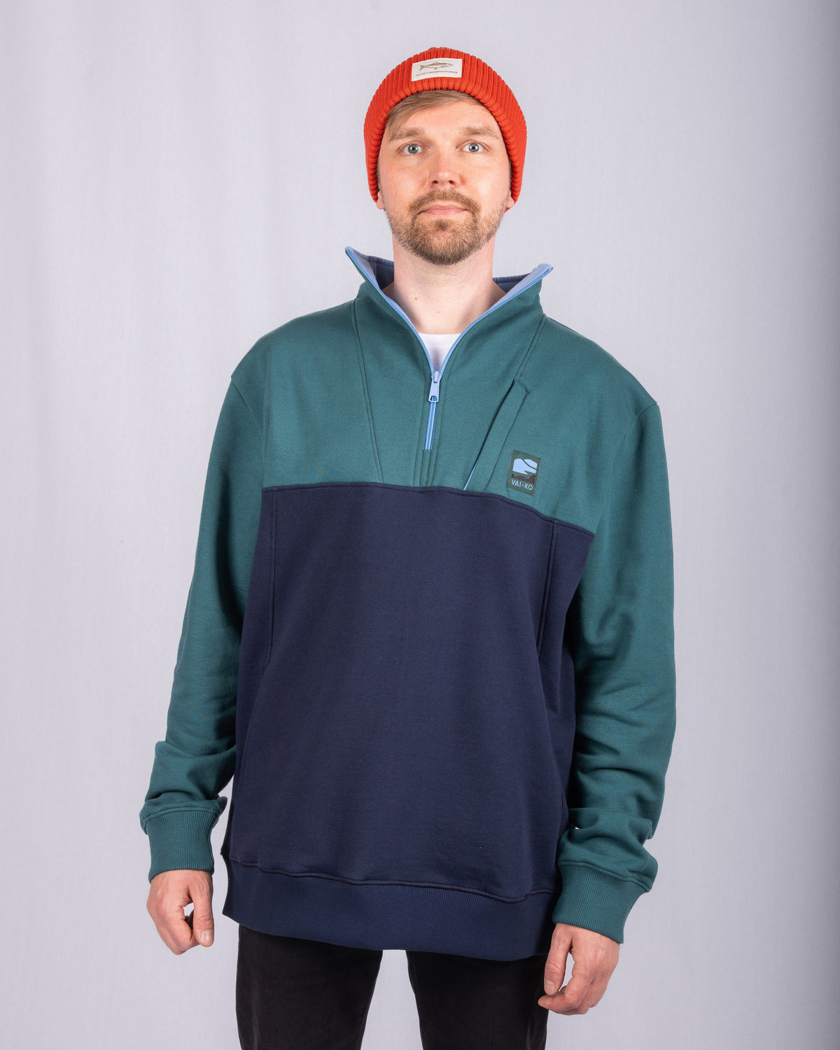 Man in a red beanie and blue-green pullover stands against a plain light gray background.