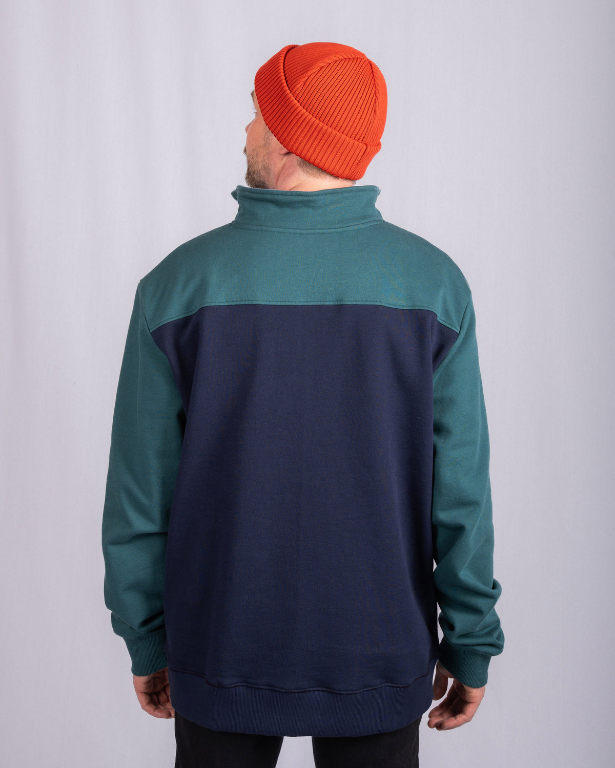 A person in a red beanie and teal-navy jacket stands facing away from the camera against a plain background.