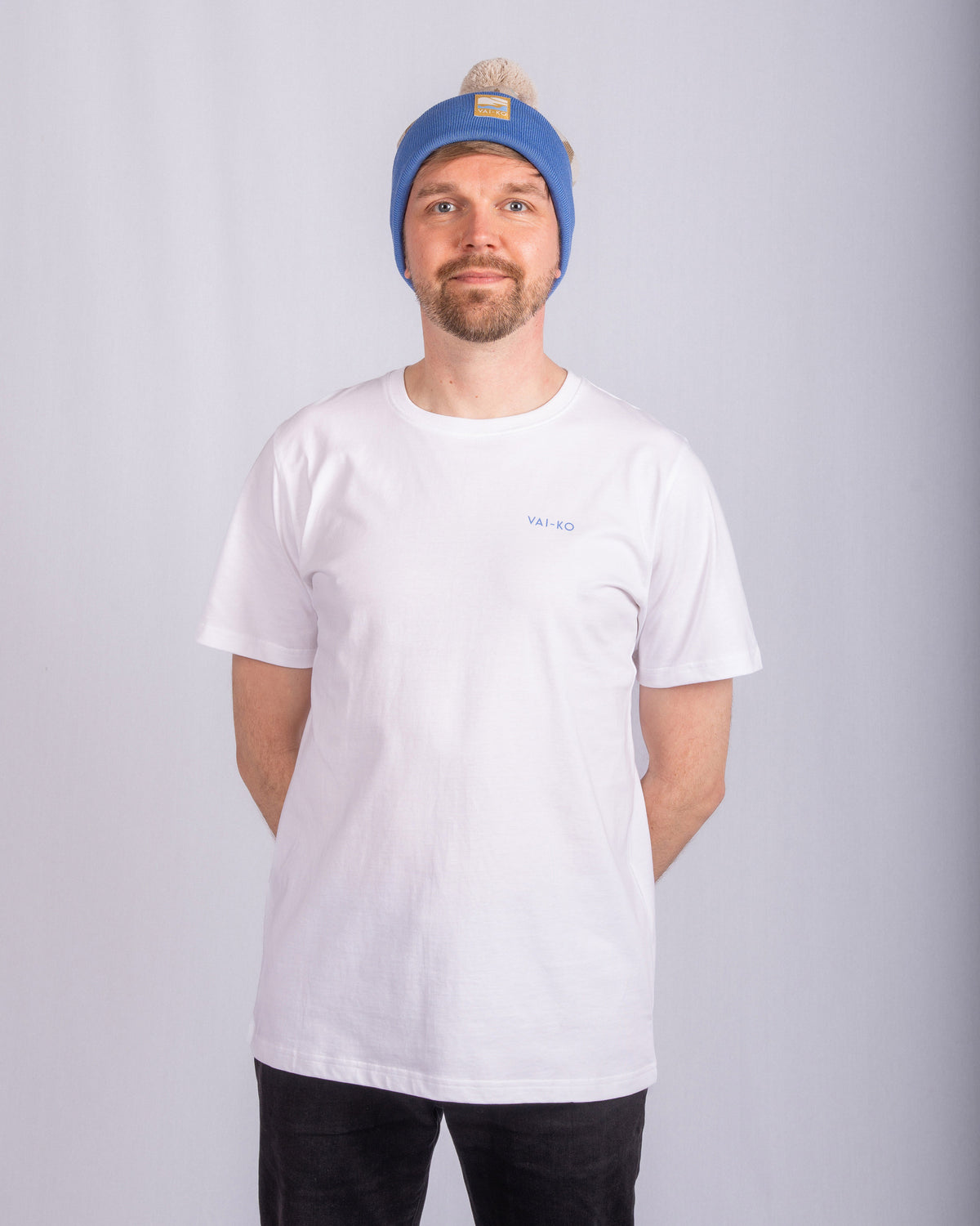 Man in a blue beanie and white t-shirt stands with hands behind his back against a plain background.
