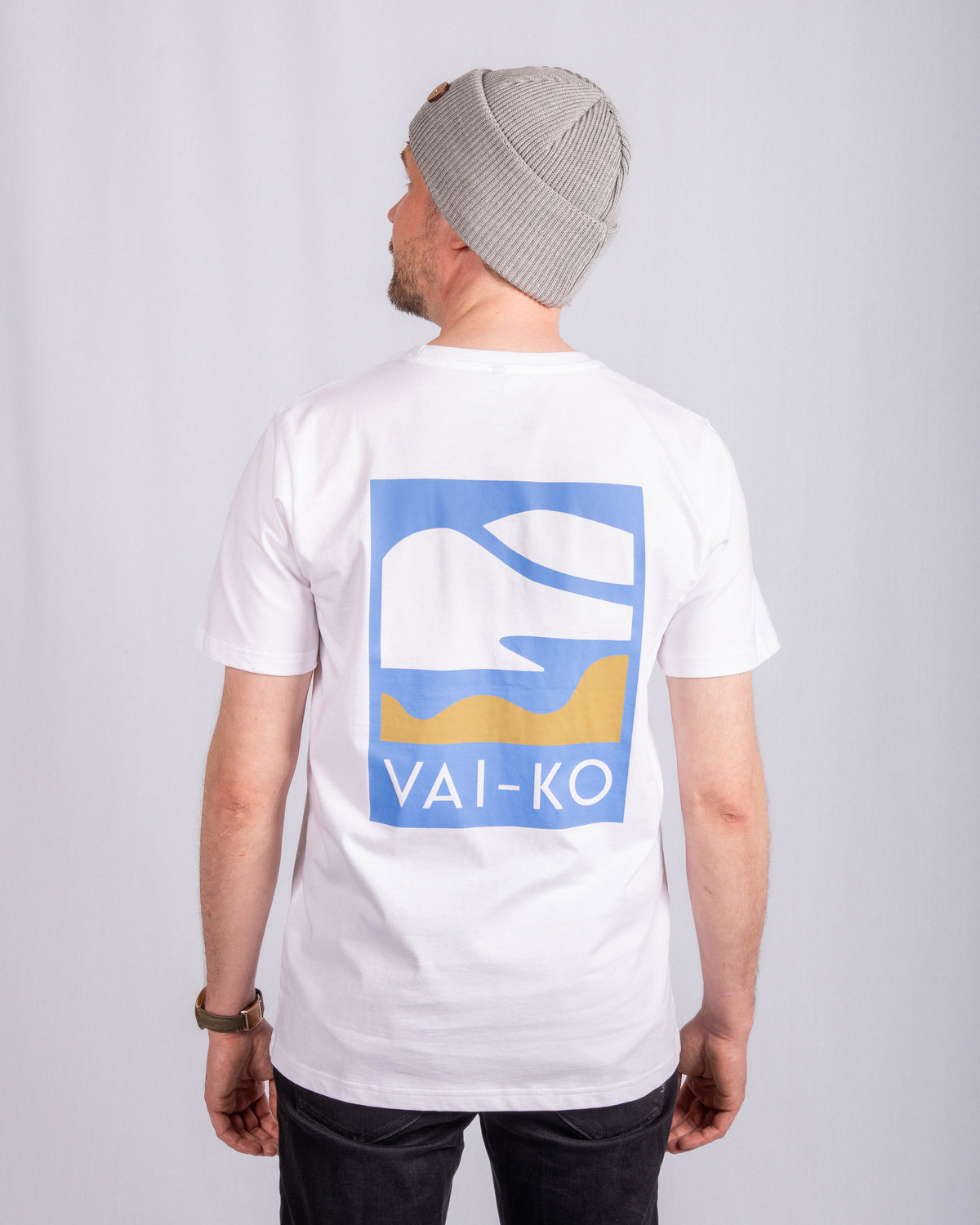 Man in gray beanie and white VAI-KO t-shirt stands facing away, logo with abstract shapes on back.