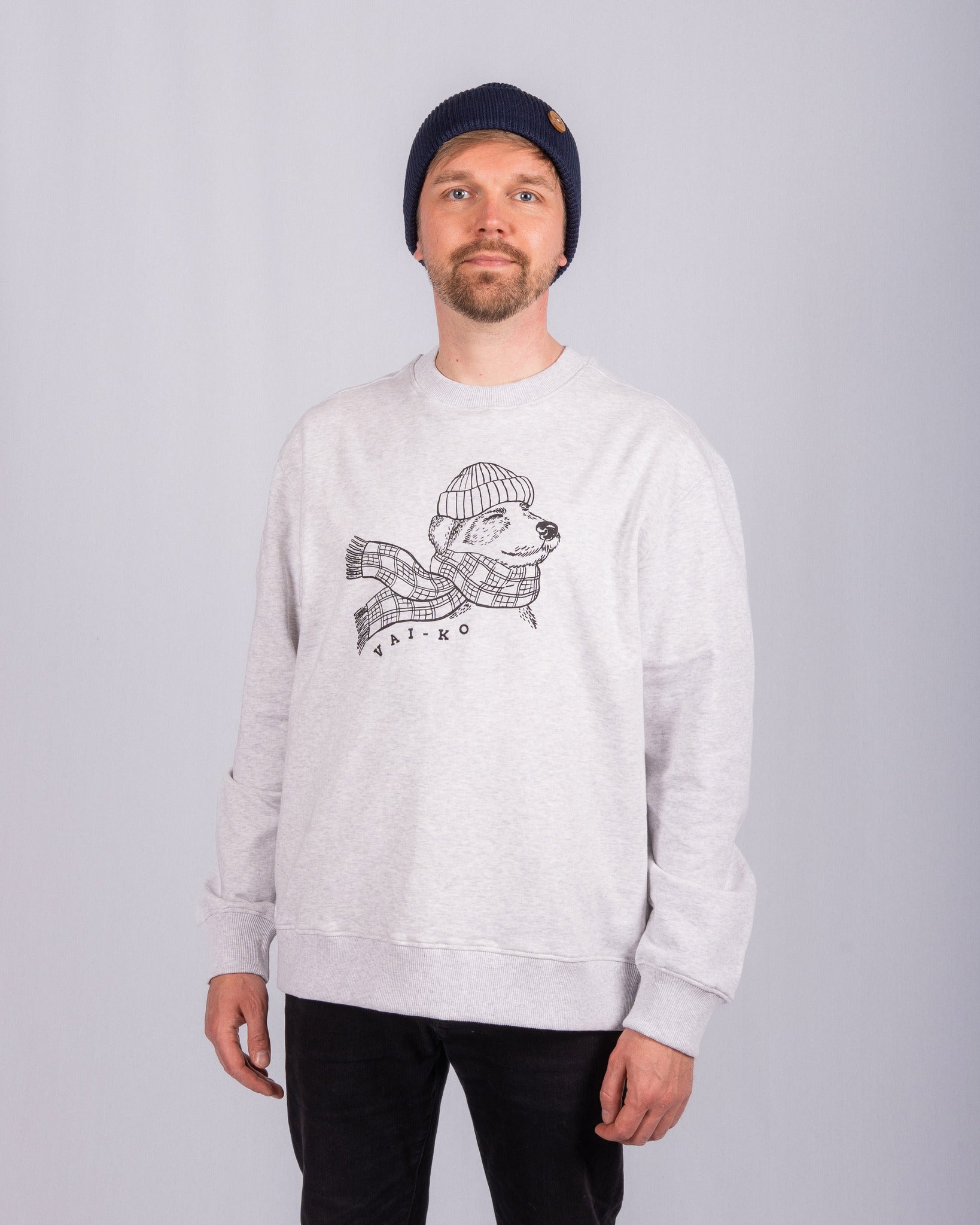 Man in beanie and light gray sweatshirt with bear and scarf graphic, standing against a plain background.