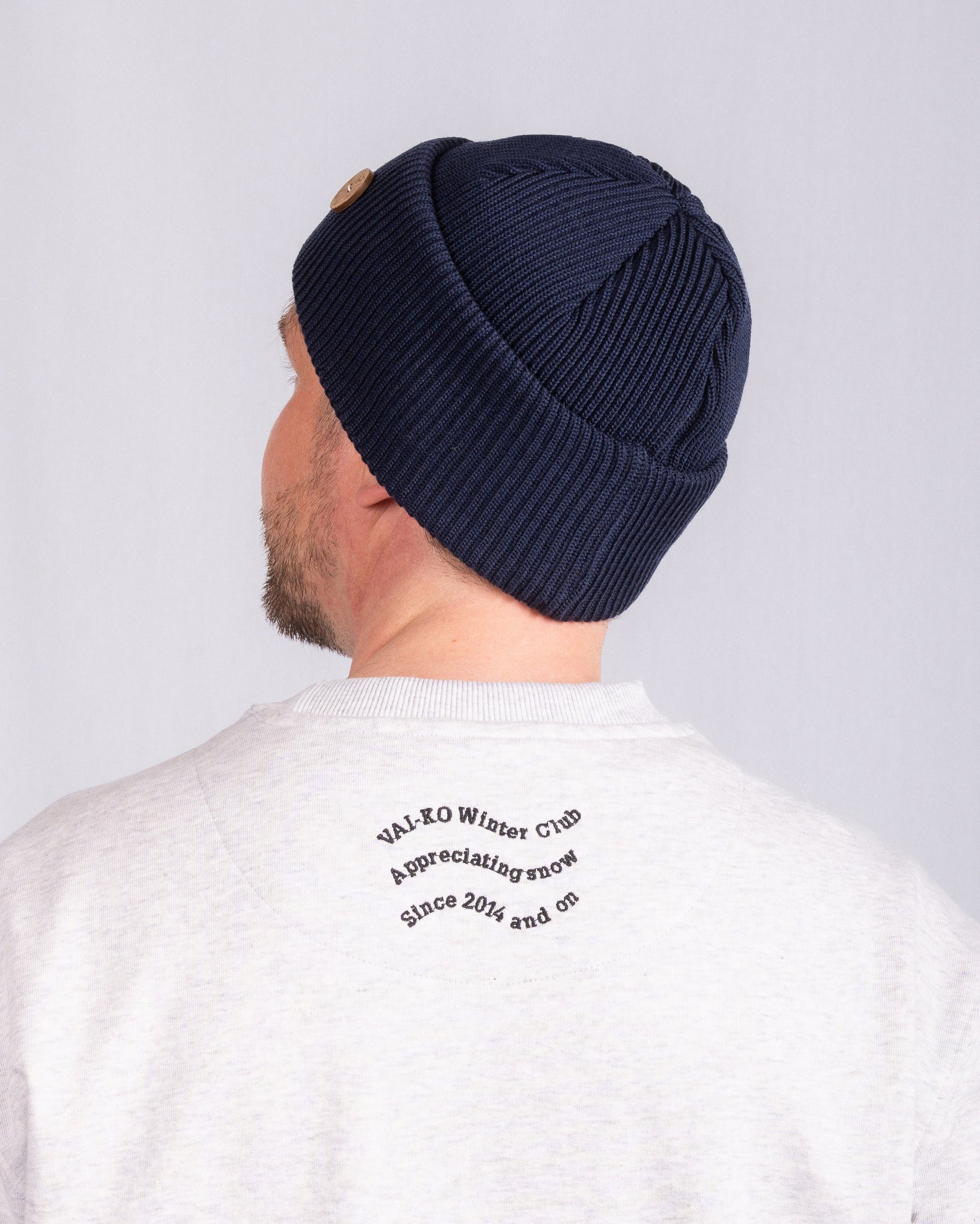 Man in navy knit beanie and white sweatshirt with printed text on the back, facing away from the camera.