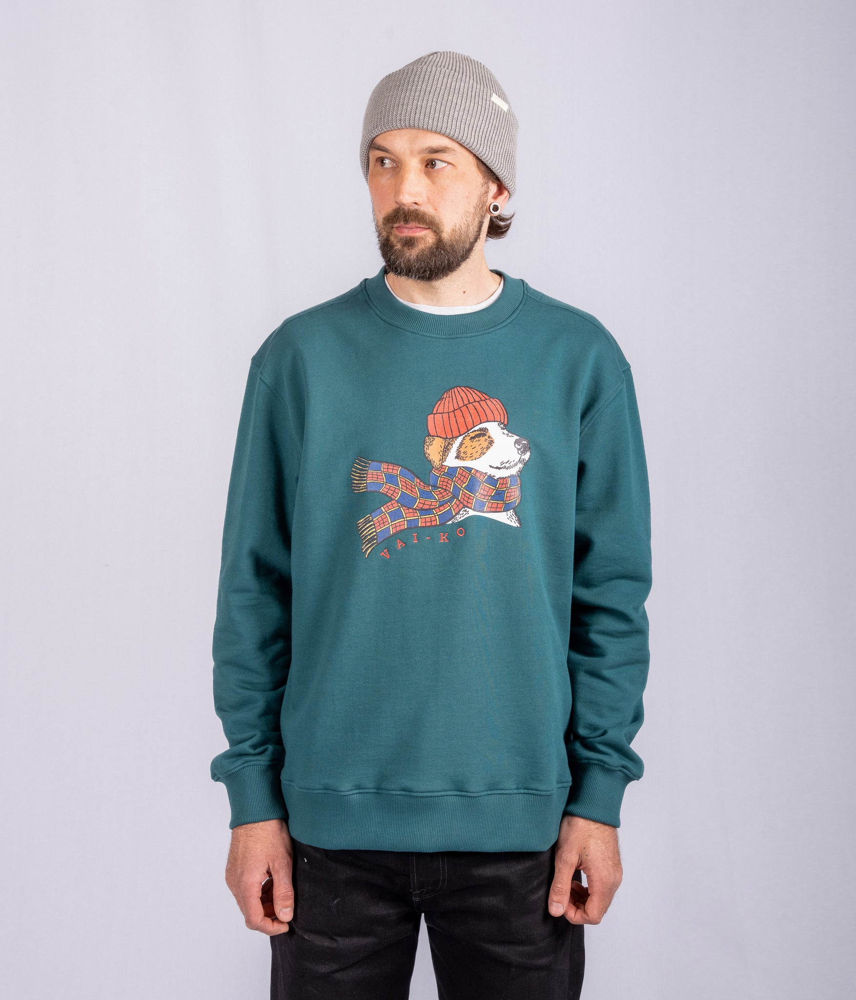 Man in green sweatshirt with dog graphic, gray beanie, standing against a plain light background.