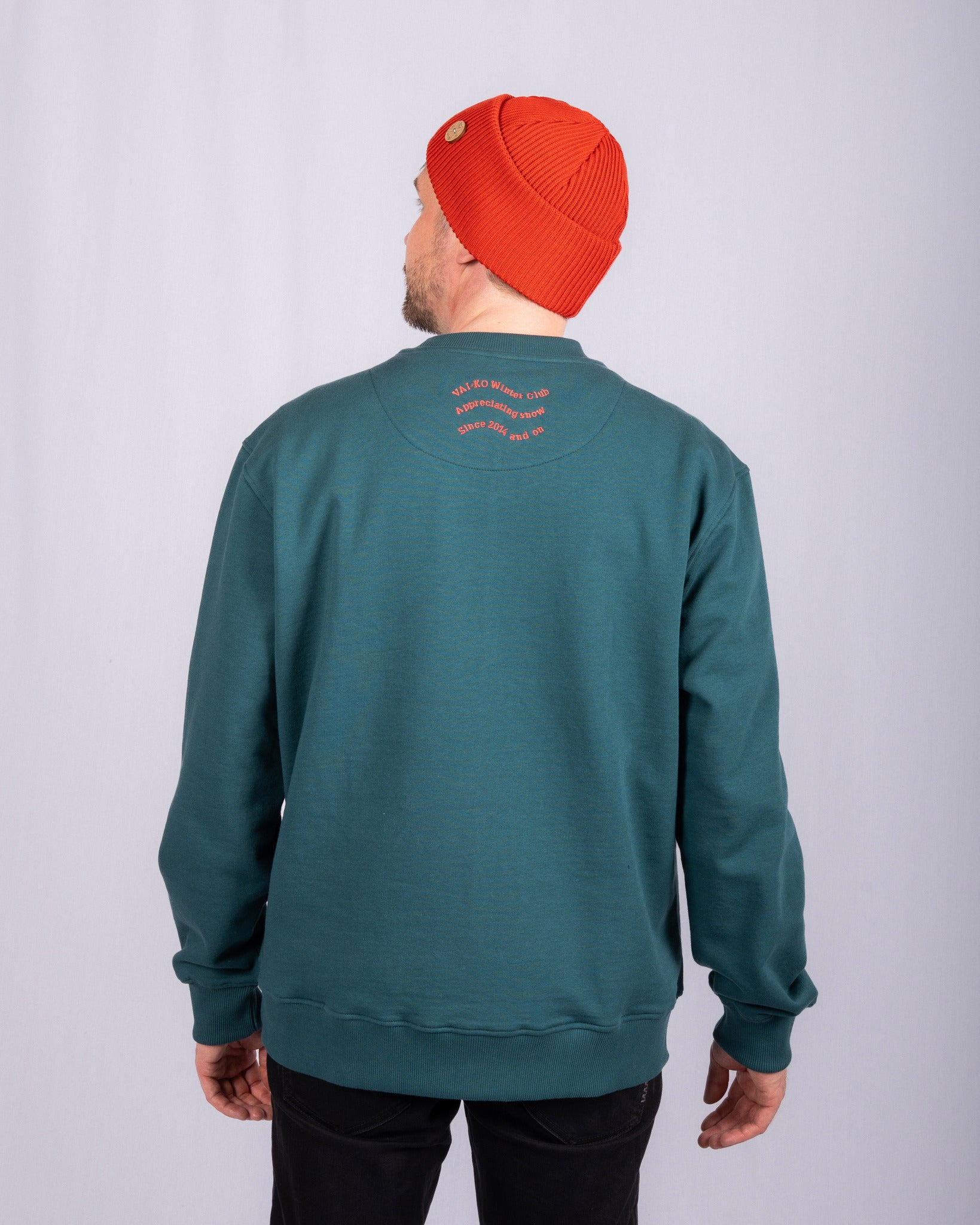 Person in a teal sweatshirt and orange beanie facing away against a plain white background.