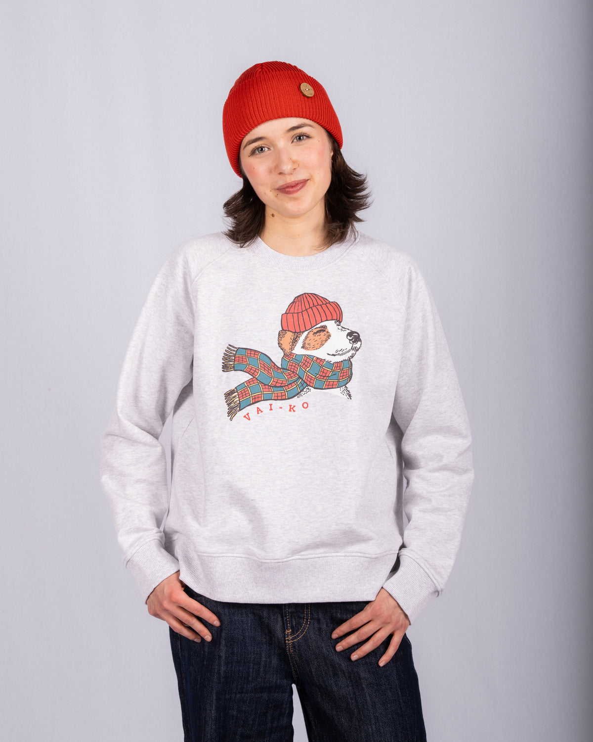 Woman in red beanie and gray sweatshirt with a dog in a scarf and hat graphic, standing and smiling.