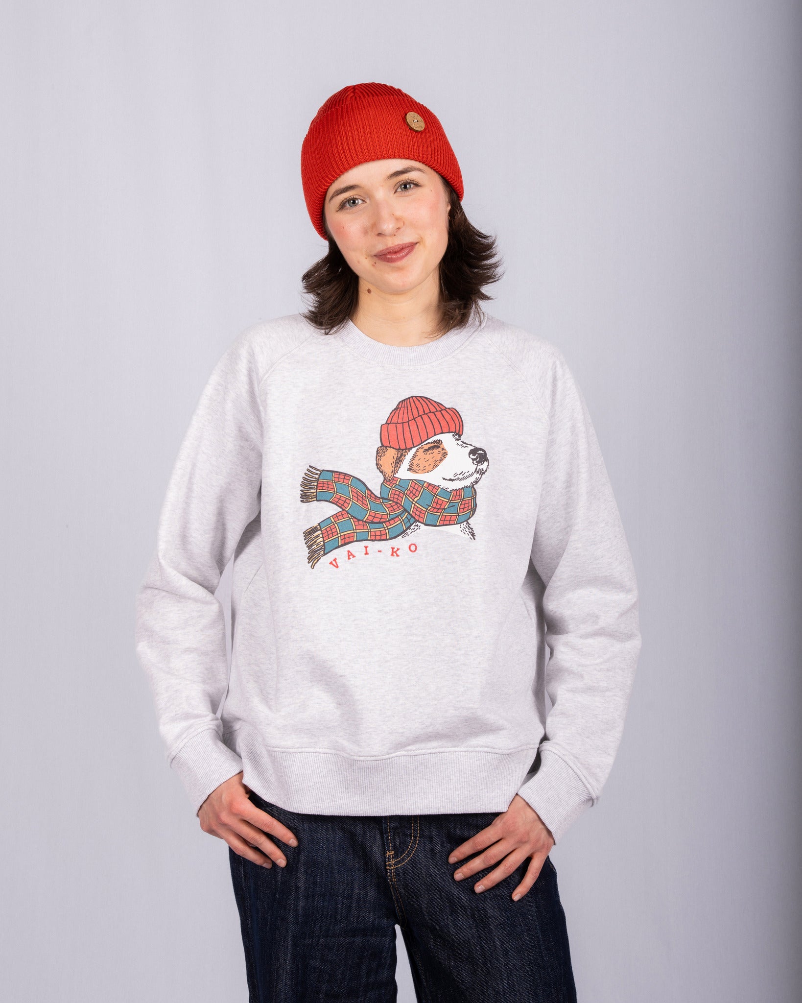 Woman in red beanie and gray sweatshirt with a dog in a scarf and hat graphic, standing and smiling.