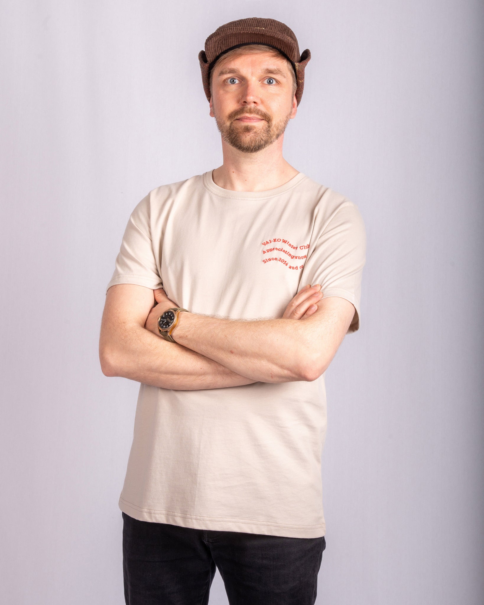 Man in a beige t-shirt and brown cap stands with arms crossed, looking at the camera, on a light background.