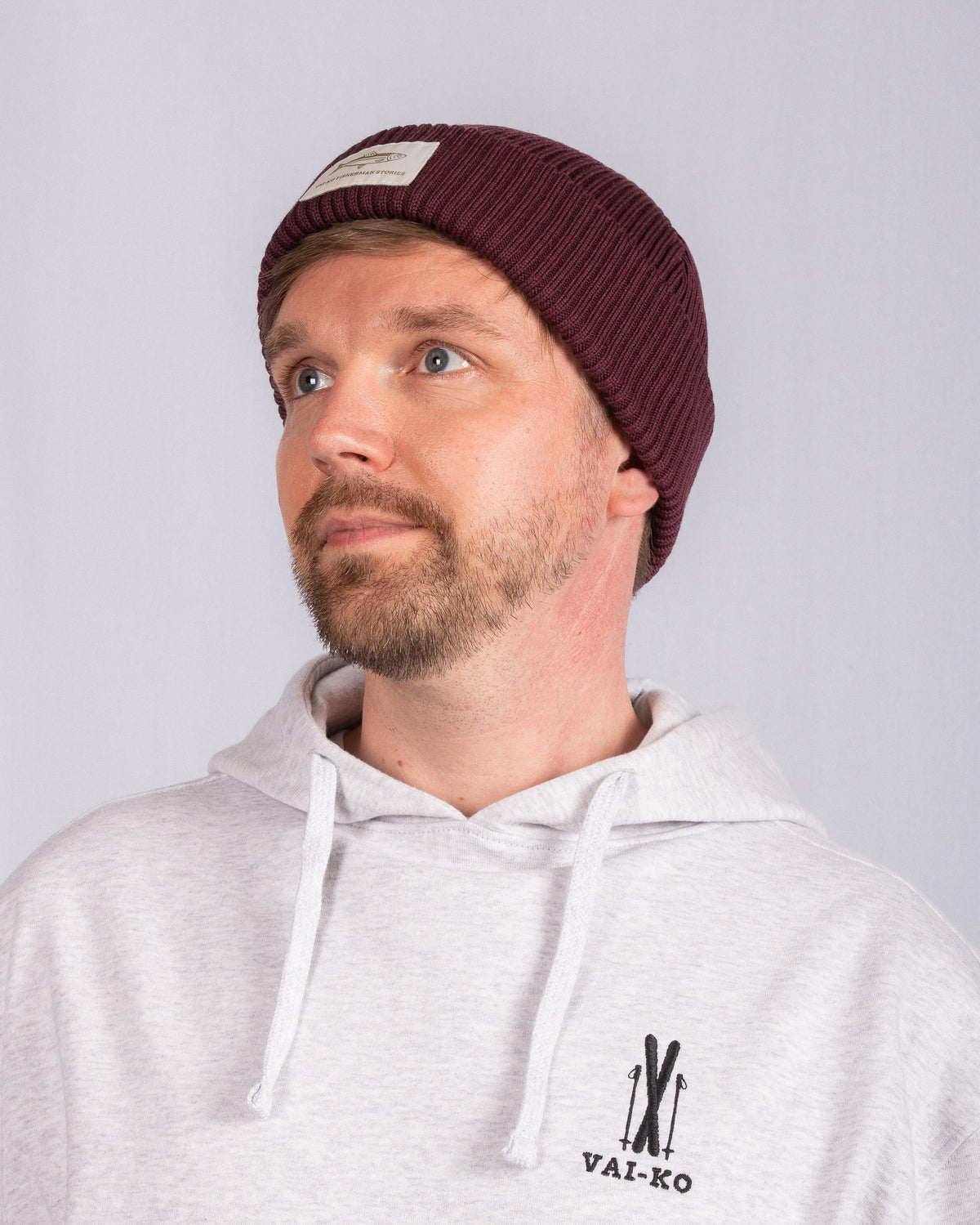 Man with a trimmed beard in a maroon beanie and gray hoodie looking up against a plain background.