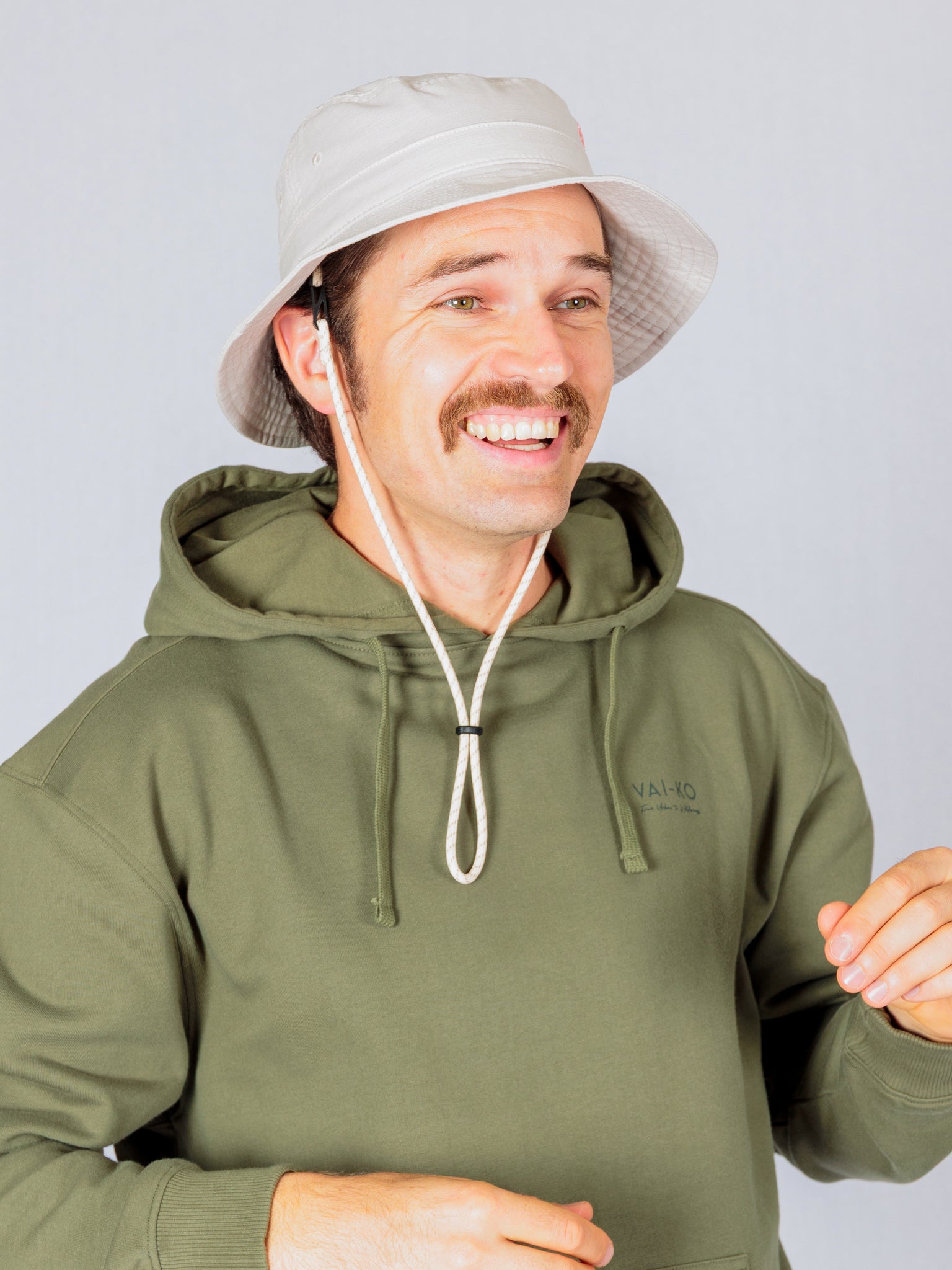 Man in a green hoodie and bucket hat smiling against a light gray background.