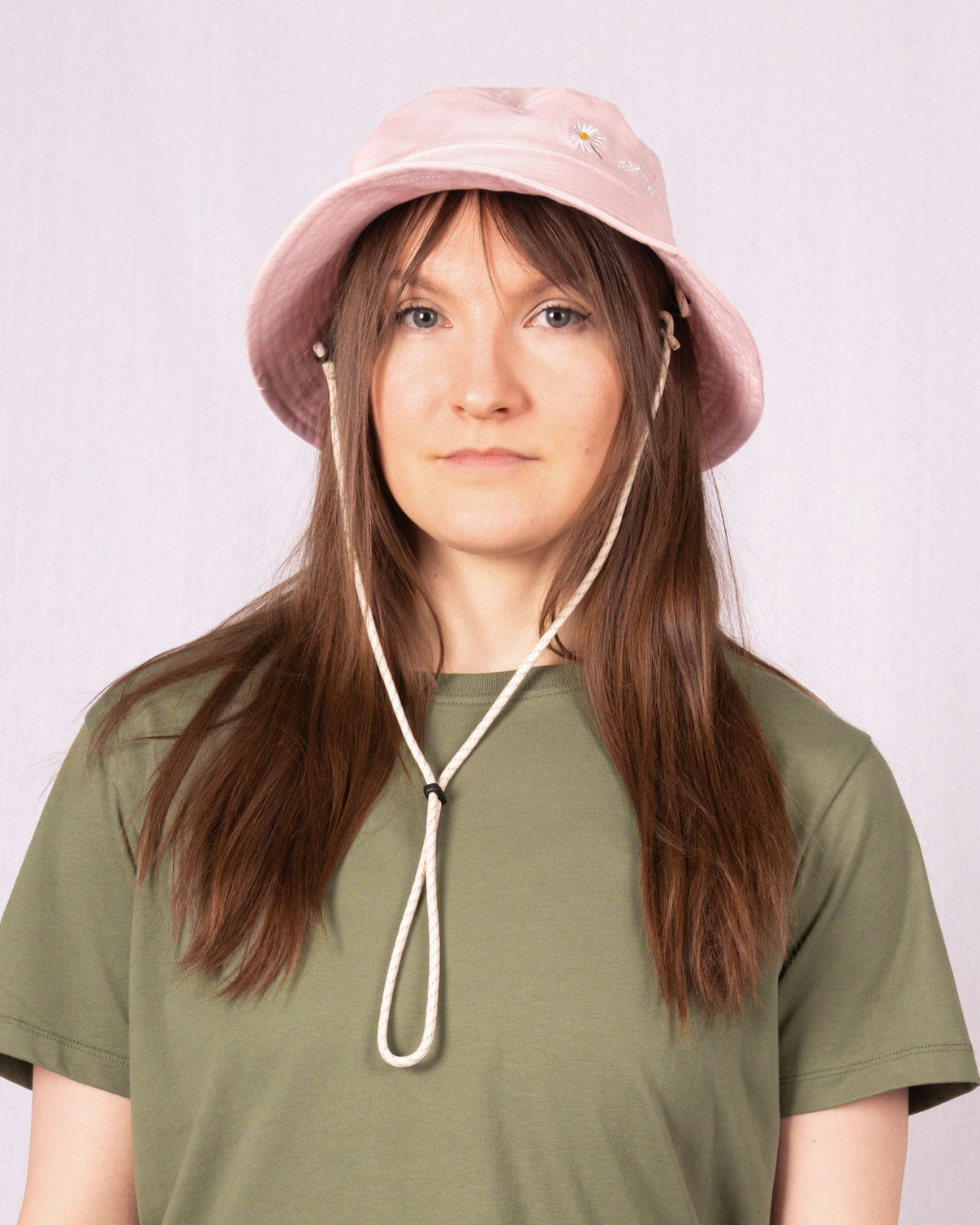 A person with long brown hair wears a pink bucket hat and olive green t-shirt, standing against a light background.
