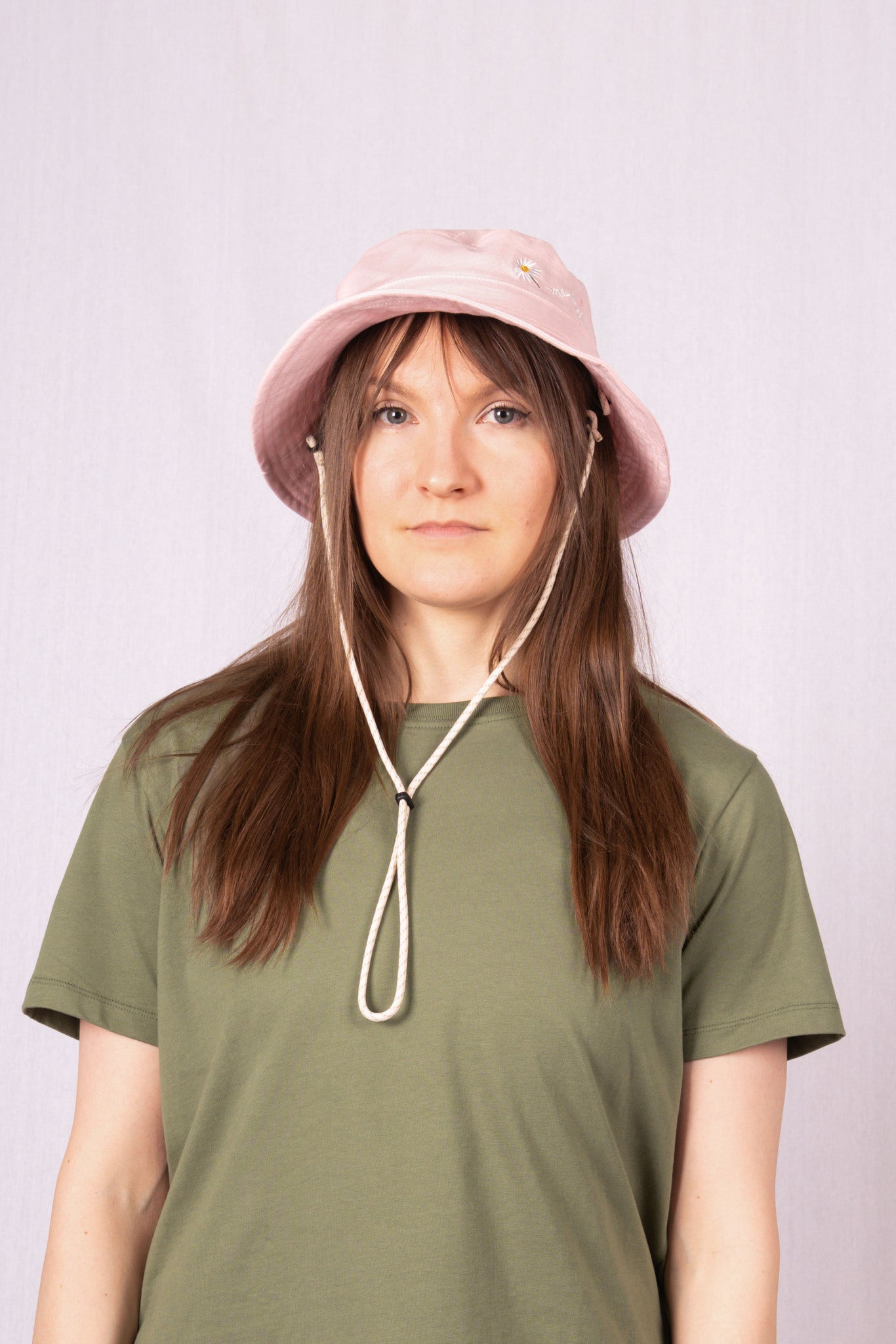 A person with long brown hair wears a pink bucket hat and olive green t-shirt, standing against a light background.