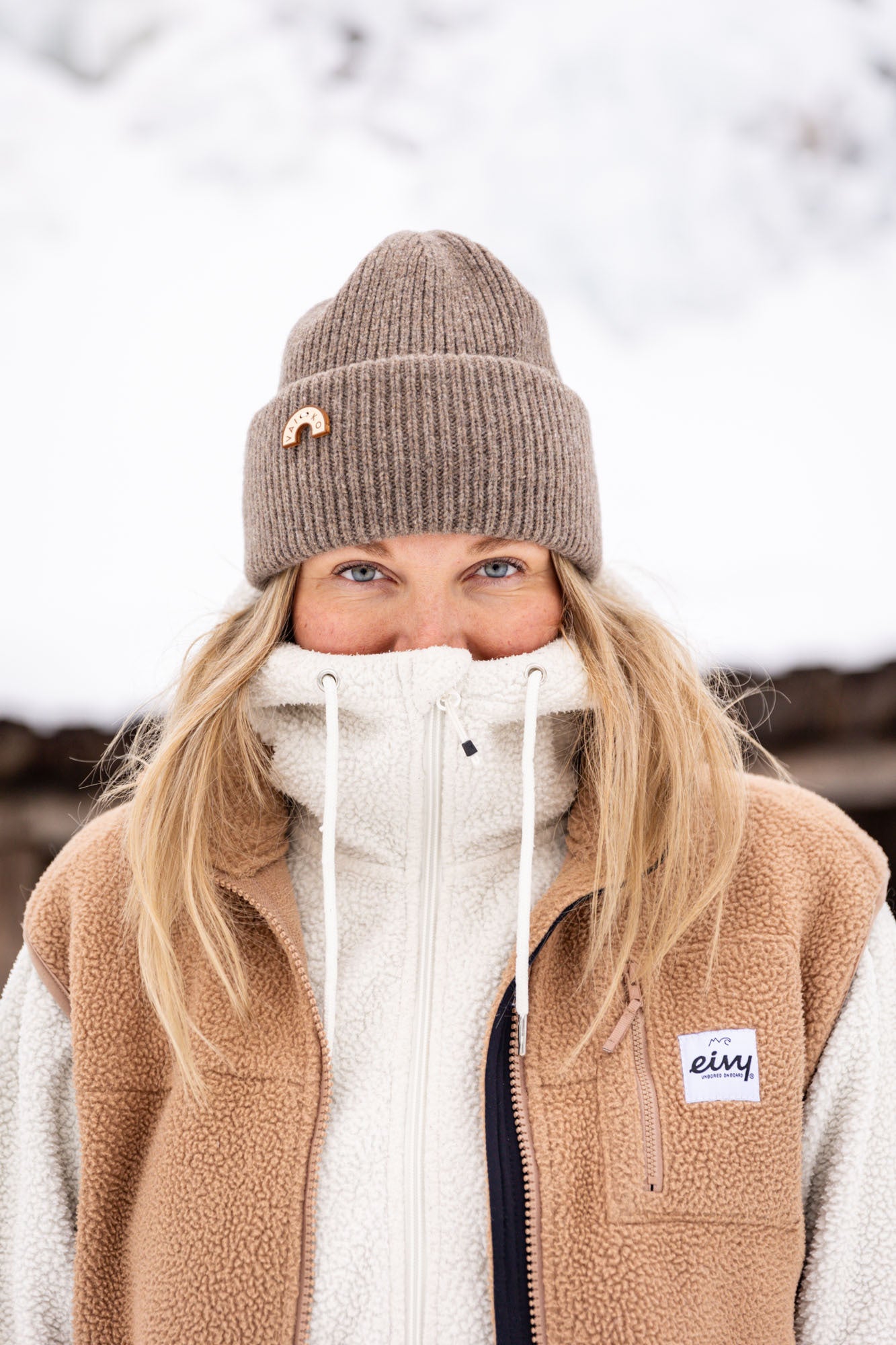 Woman in a brown beanie and fleece jacket outdoors, zipped up to her nose, with snow in the background.