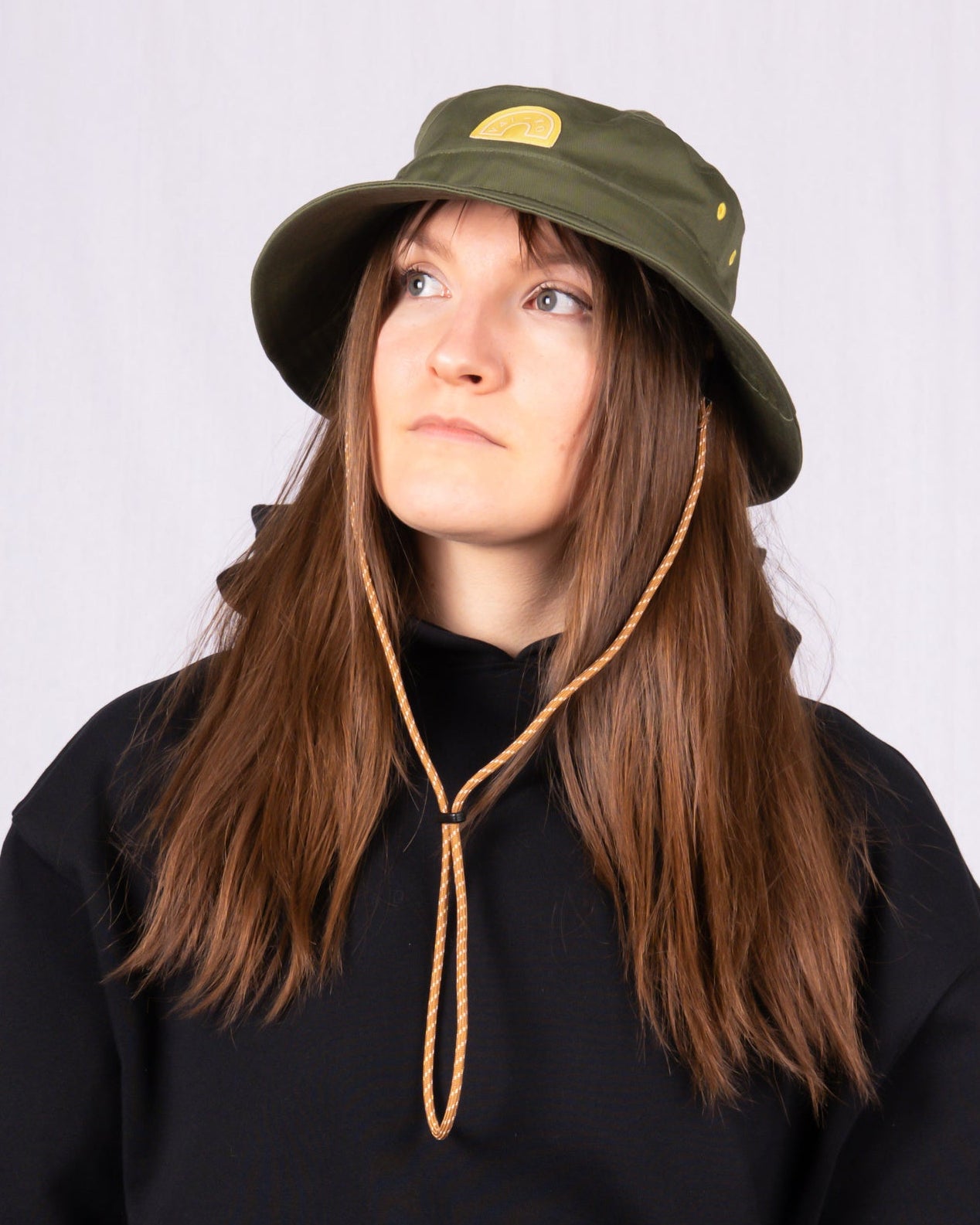 Woman in a green bucket hat and black hoodie looking up, with long brown hair and a neutral expression.