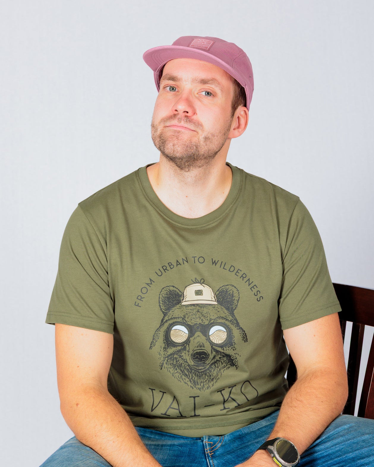 Man in pink cap and green t-shirt with bear graphic sits on a chair, smiling slightly at the camera.