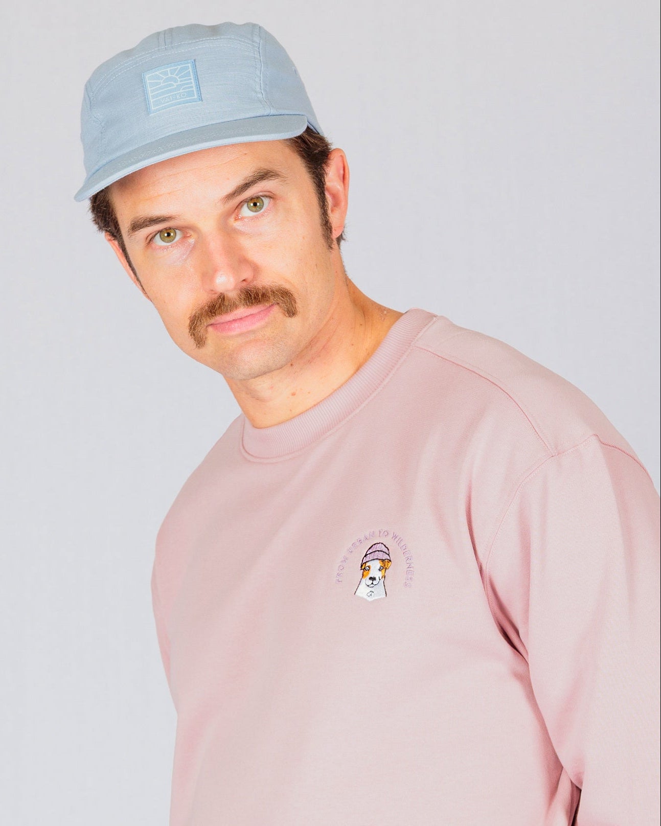 Man with a mustache wearing a light blue cap and pink sweatshirt, standing against a plain background.