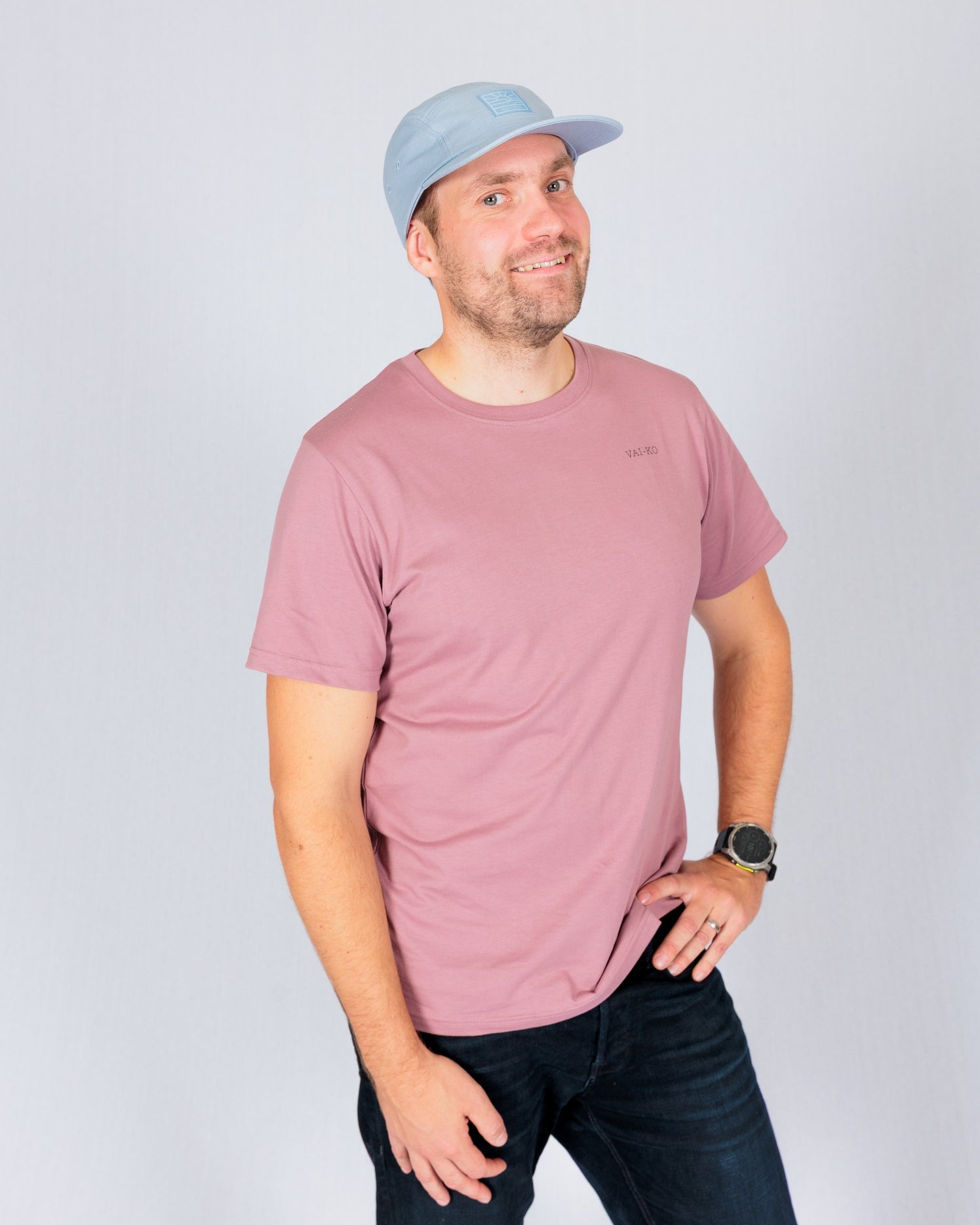 Man in a pink t-shirt and light blue cap smiling, standing with one hand on hip against a plain background.
