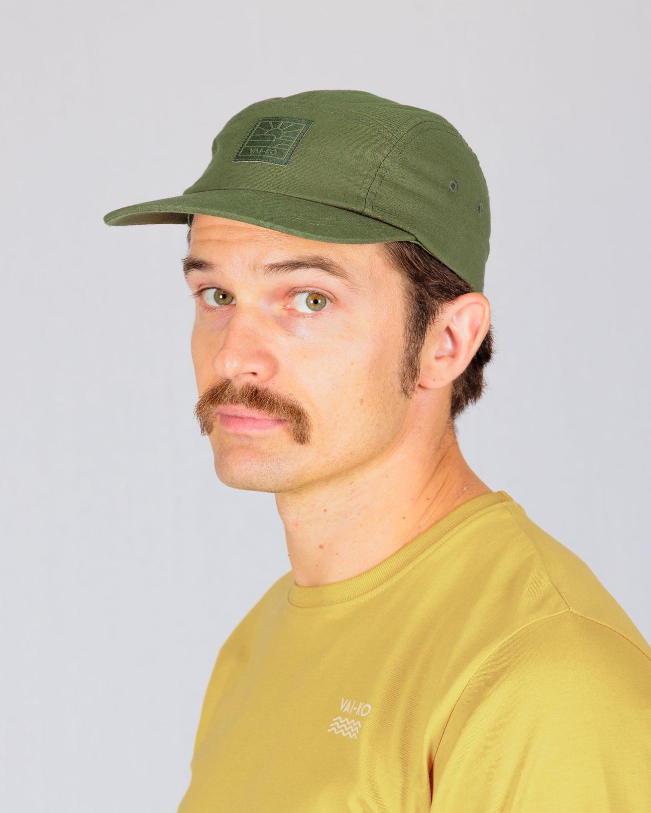 Man with a mustache wearing a green cap and yellow shirt, looking to the side against a light background.