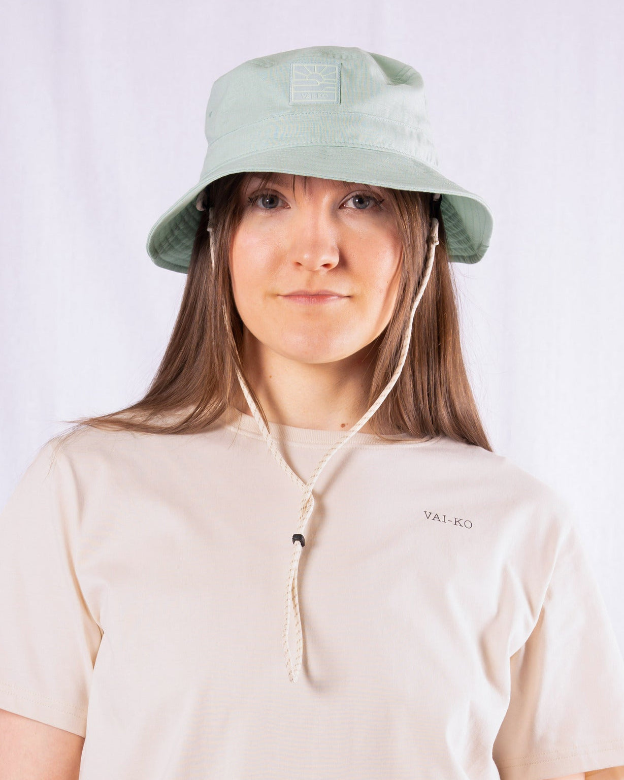 A person wearing a light green bucket hat and a beige T-shirt with the text VAI-KO on it.