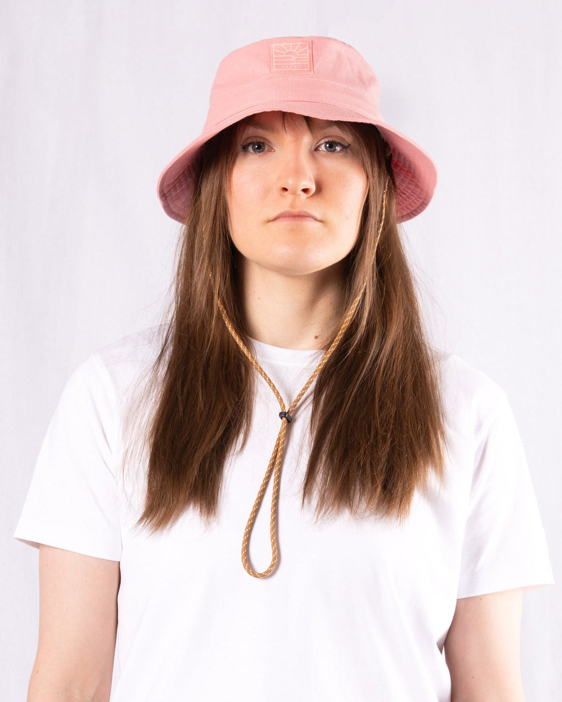 A person in a pink bucket hat and white t-shirt stands against a plain white background, looking at the camera.