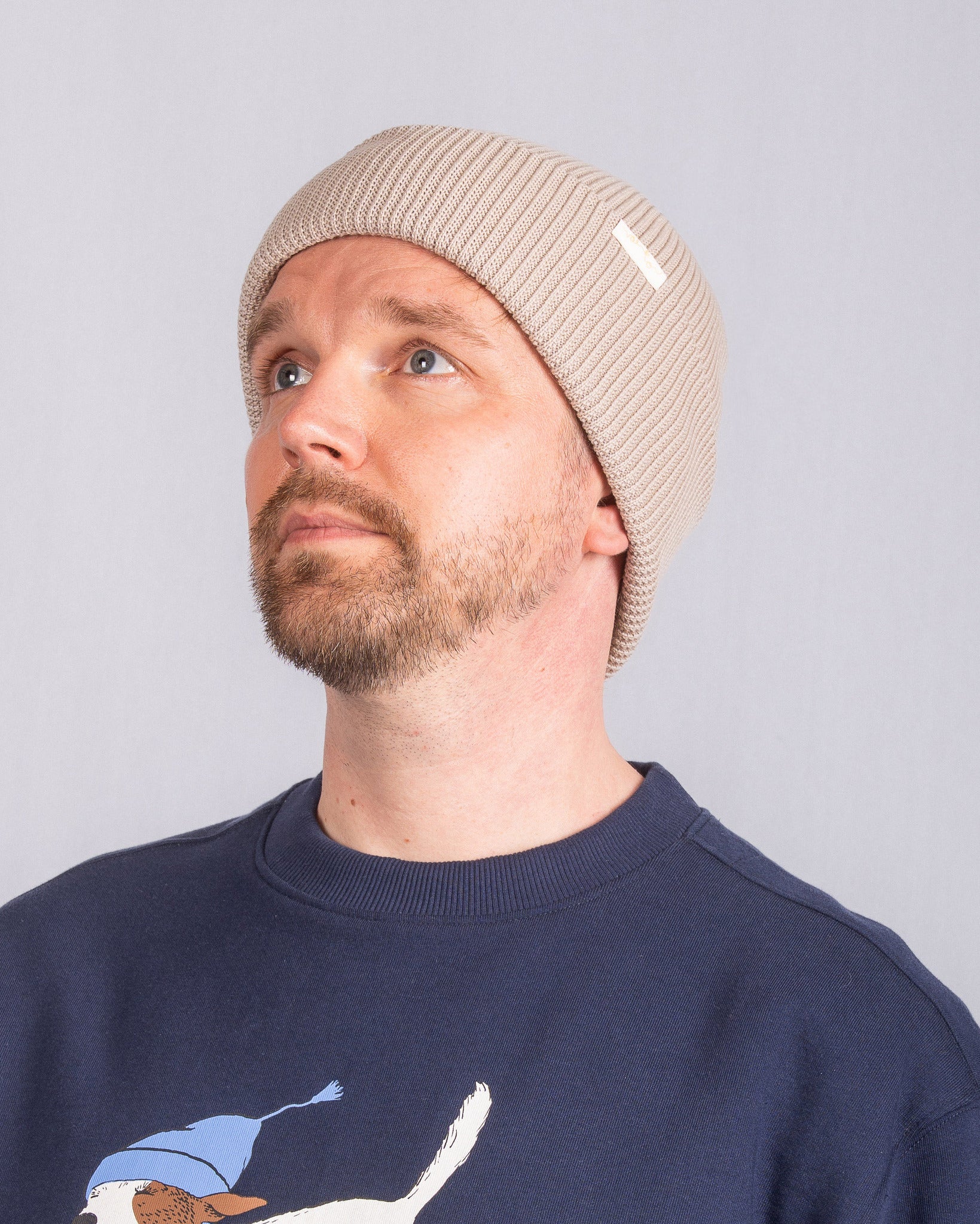 Man with a beard wearing a beige knit beanie and navy sweater, looking upwards against a plain background.