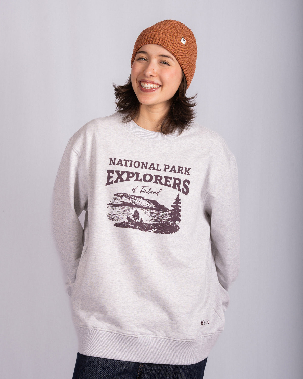 Smiling person in a tan beanie and gray National Park Explorers sweatshirt stands against a plain background.