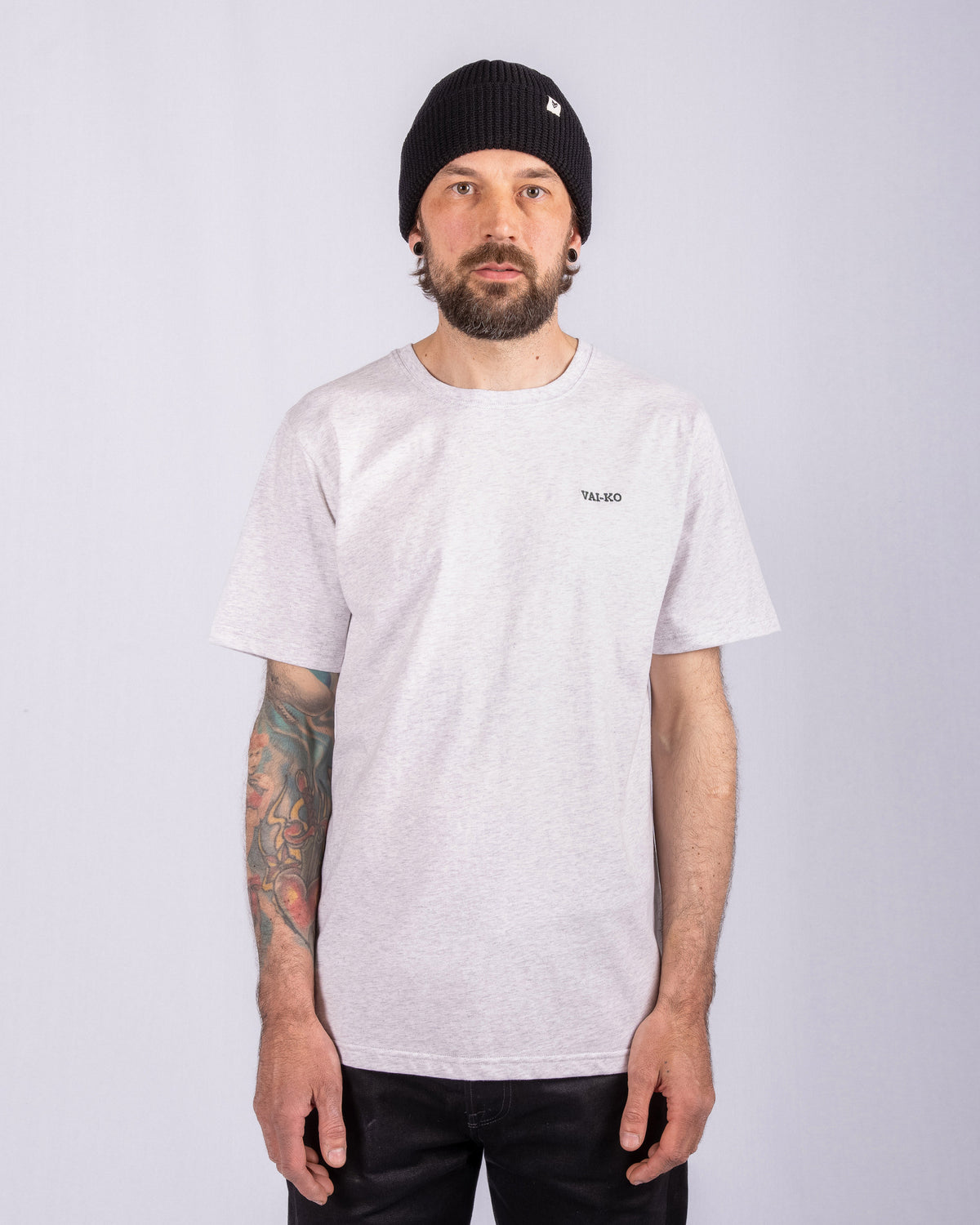Man with a beard, wearing a black beanie and light gray t-shirt, stands against a plain background.