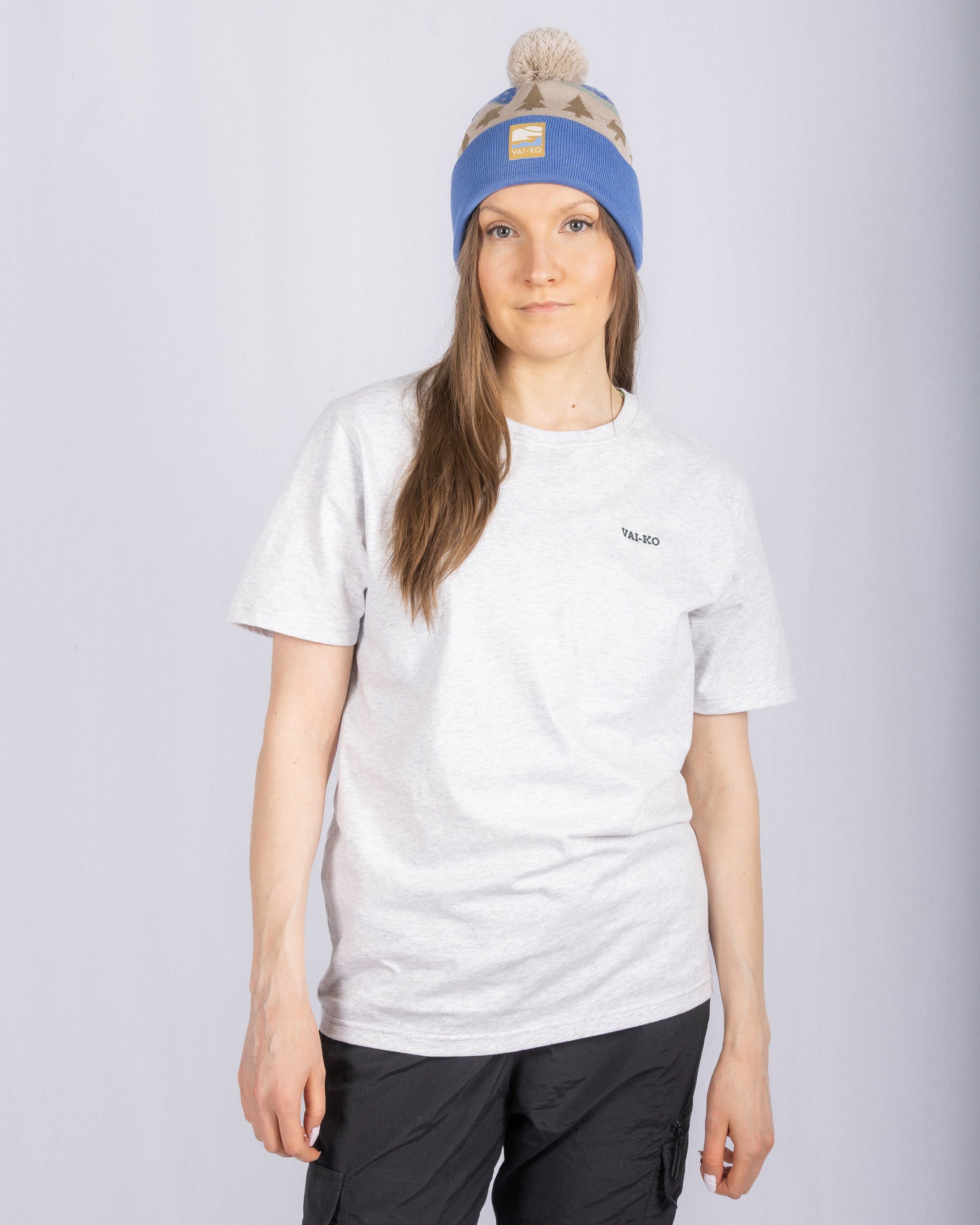 Woman in a blue and beige beanie and light gray T-shirt standing against a plain light background.