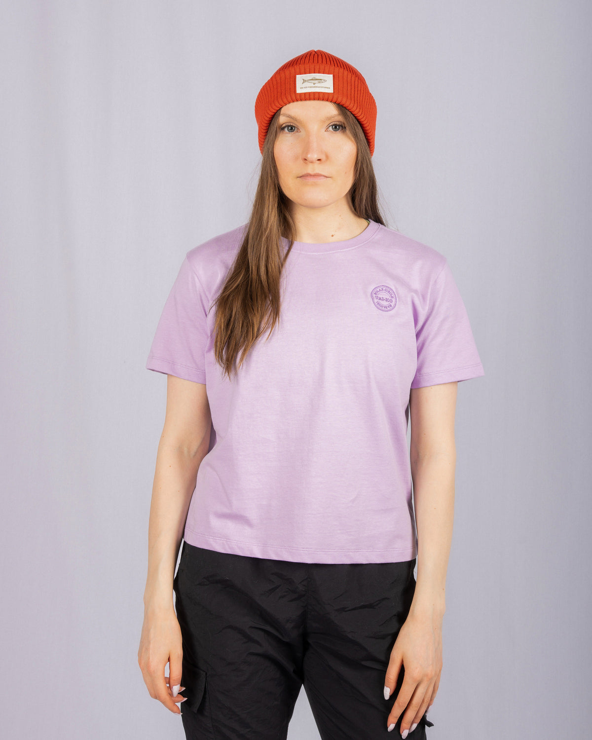 Woman in a red beanie and lavender t-shirt stands against a plain light gray background.