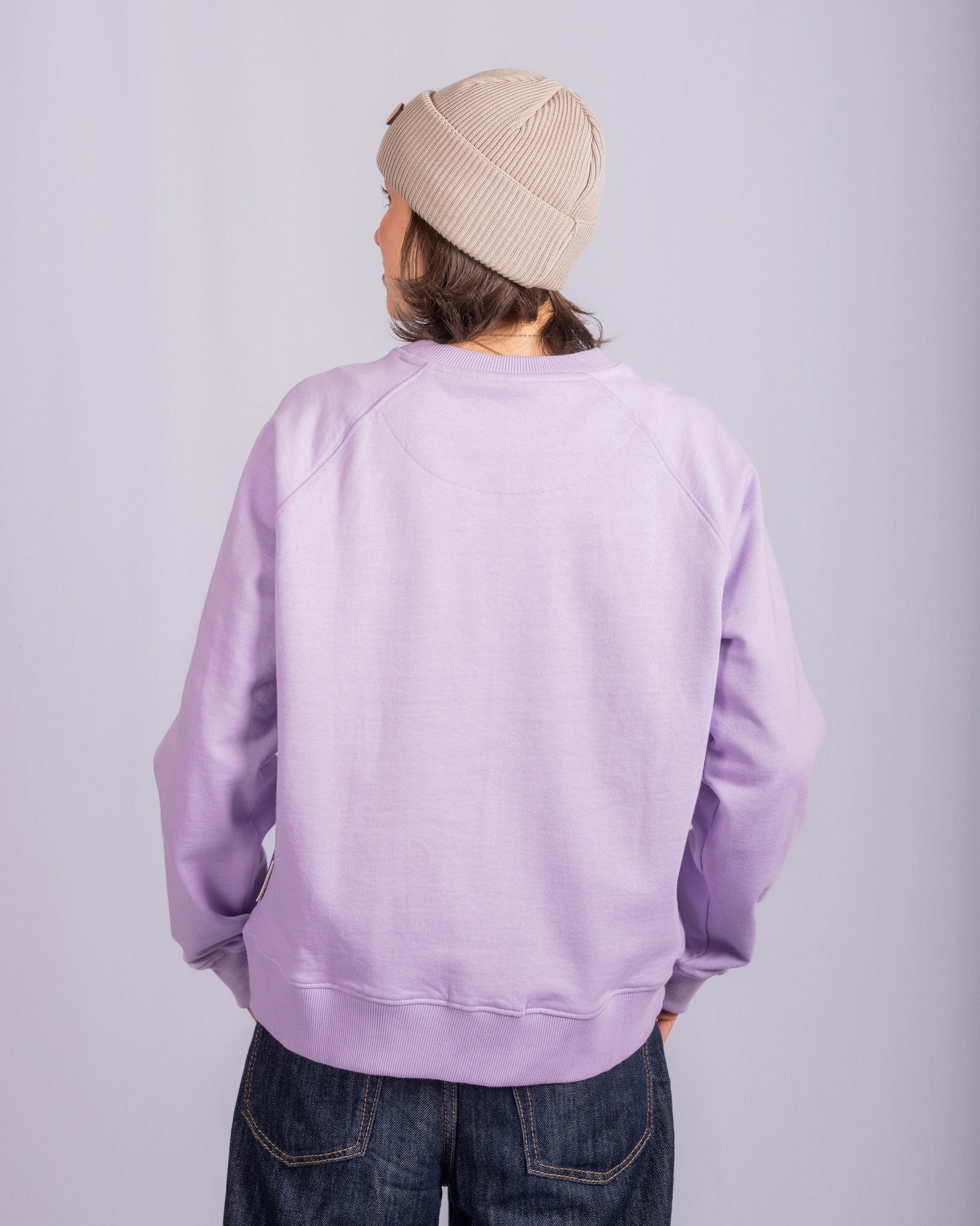 Person in a beige beanie and lilac sweatshirt stands facing away, hands on hips, against a plain background.