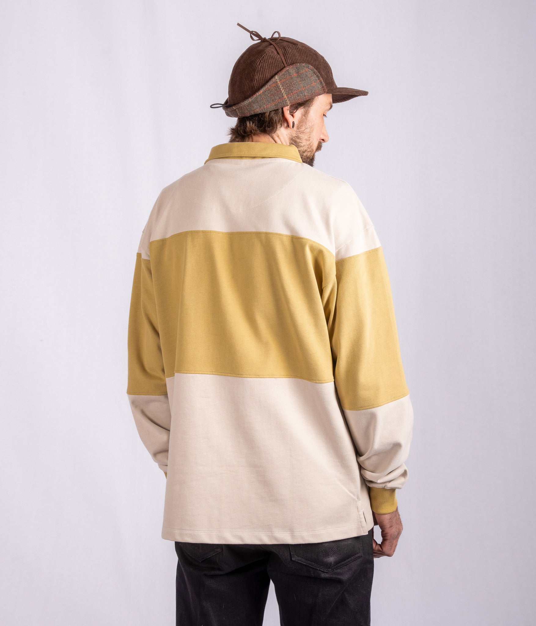 Man facing away, wearing a brown cap and a beige and mustard color-blocked sweater, with black pants.