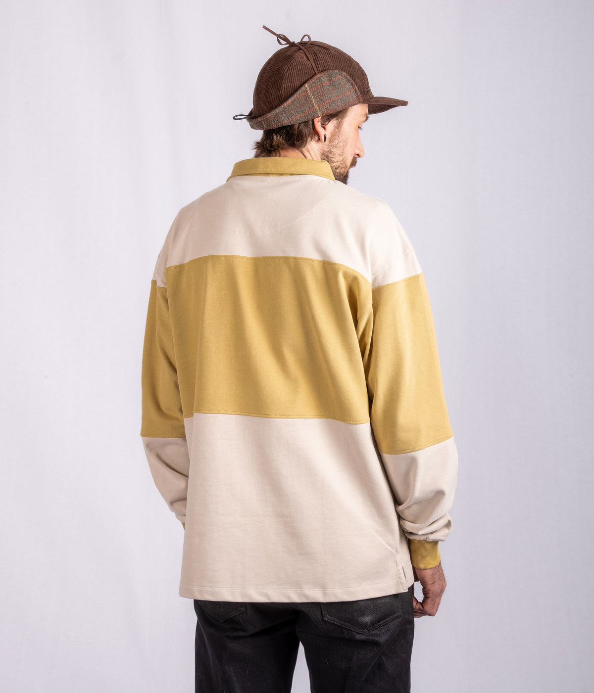 Man facing away, wearing a brown cap and a beige and mustard color-blocked sweater, with black pants.