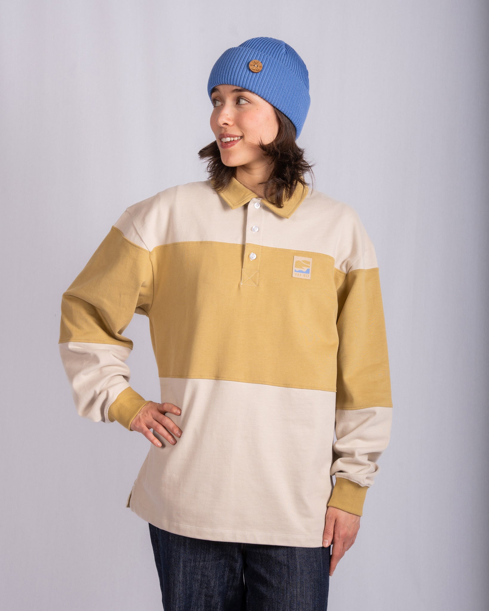 Person in blue beanie and beige-yellow striped shirt stands smiling, hand on hip, against plain backdrop.