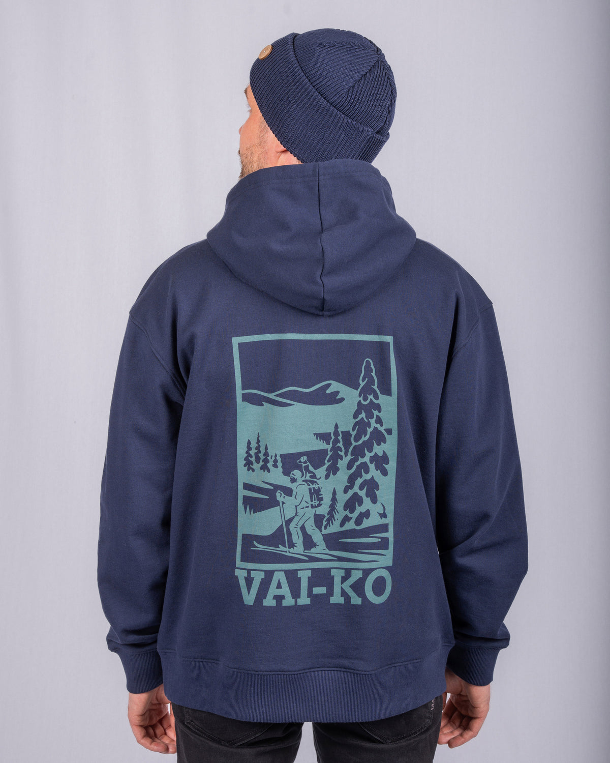Man in a navy hoodie and beanie, with a nature scene and VAI-KO logo on the back of the hoodie.