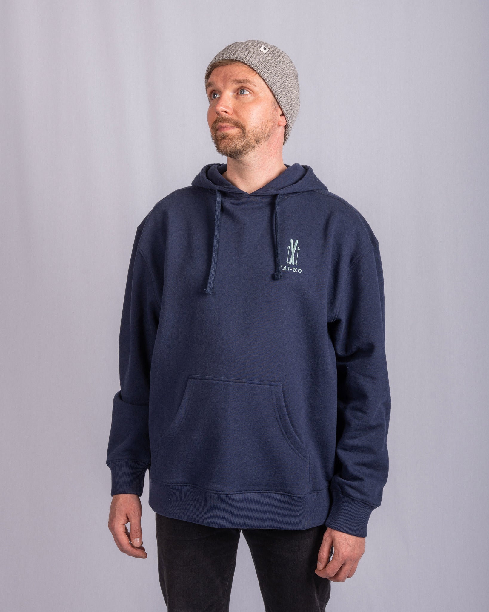 Man in a gray beanie and navy hoodie standing against a plain light gray background, looking upward.