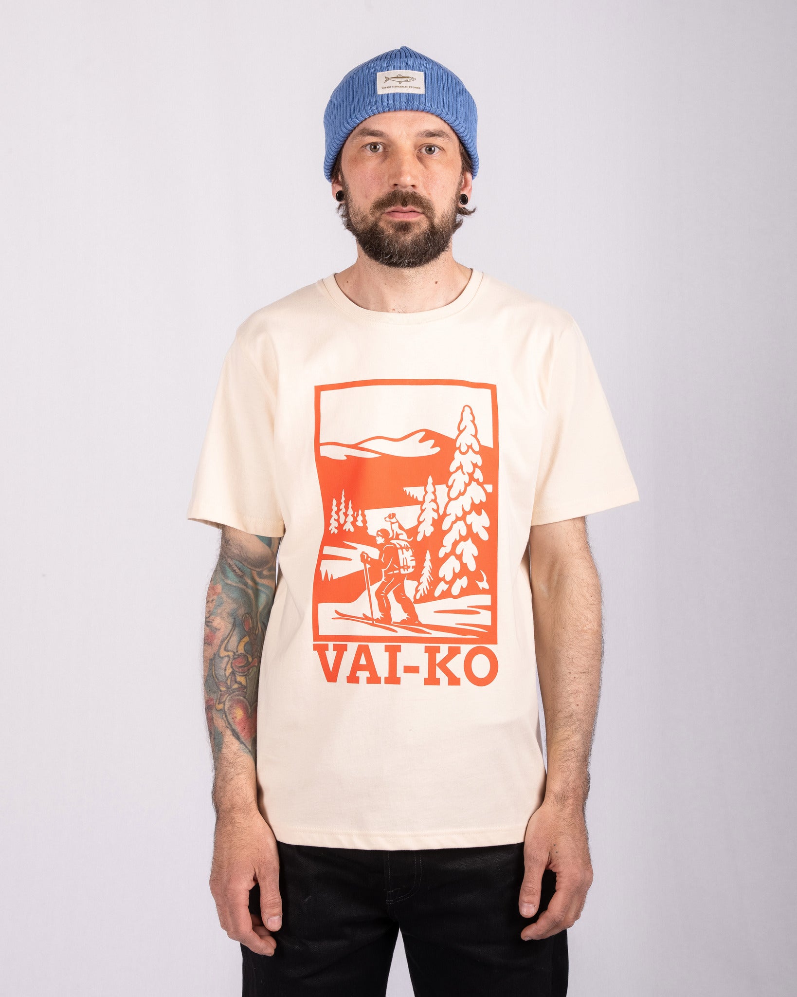 Man in blue beanie and printed t-shirt with orange mountain scene, standing against a plain background.