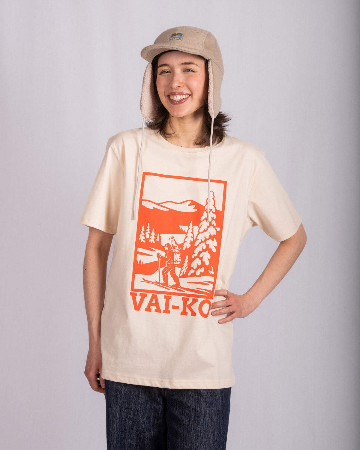 Smiling person in a beige hat and graphic T-shirt with VAI-KO text stands against a plain background.