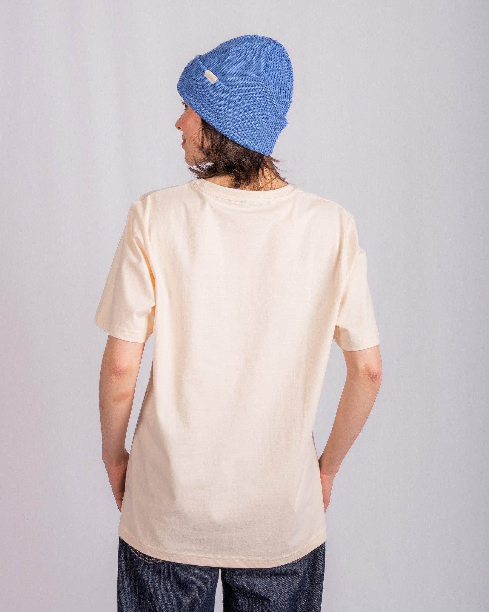Person in a blue beanie and cream t-shirt stands facing away against a plain light background.
