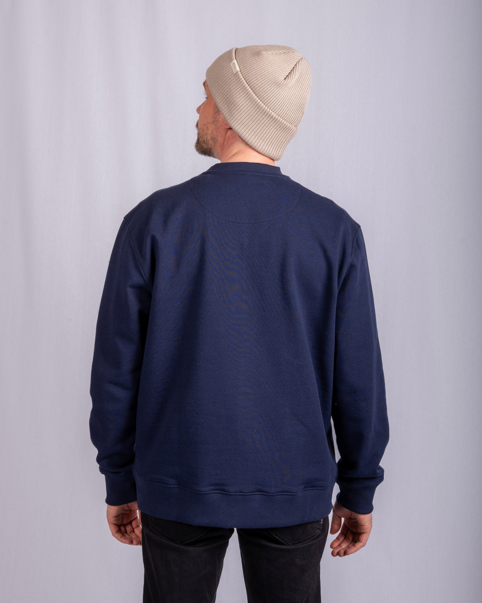 Man in beige beanie and navy blue sweatshirt stands facing away against light gray background.