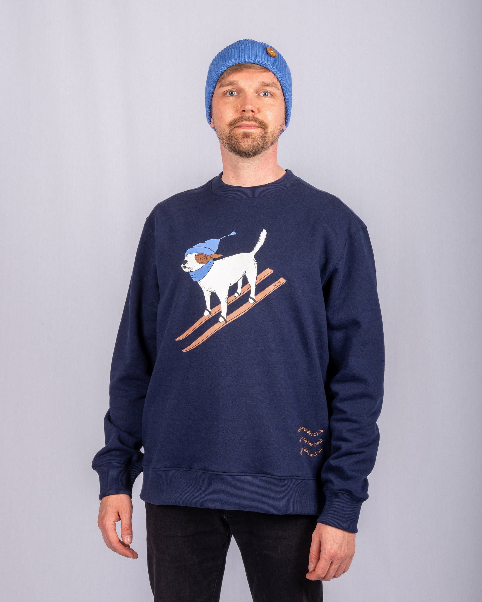 Man in a blue beanie and navy sweatshirt with a skiing dog graphic stands against a plain gray background.