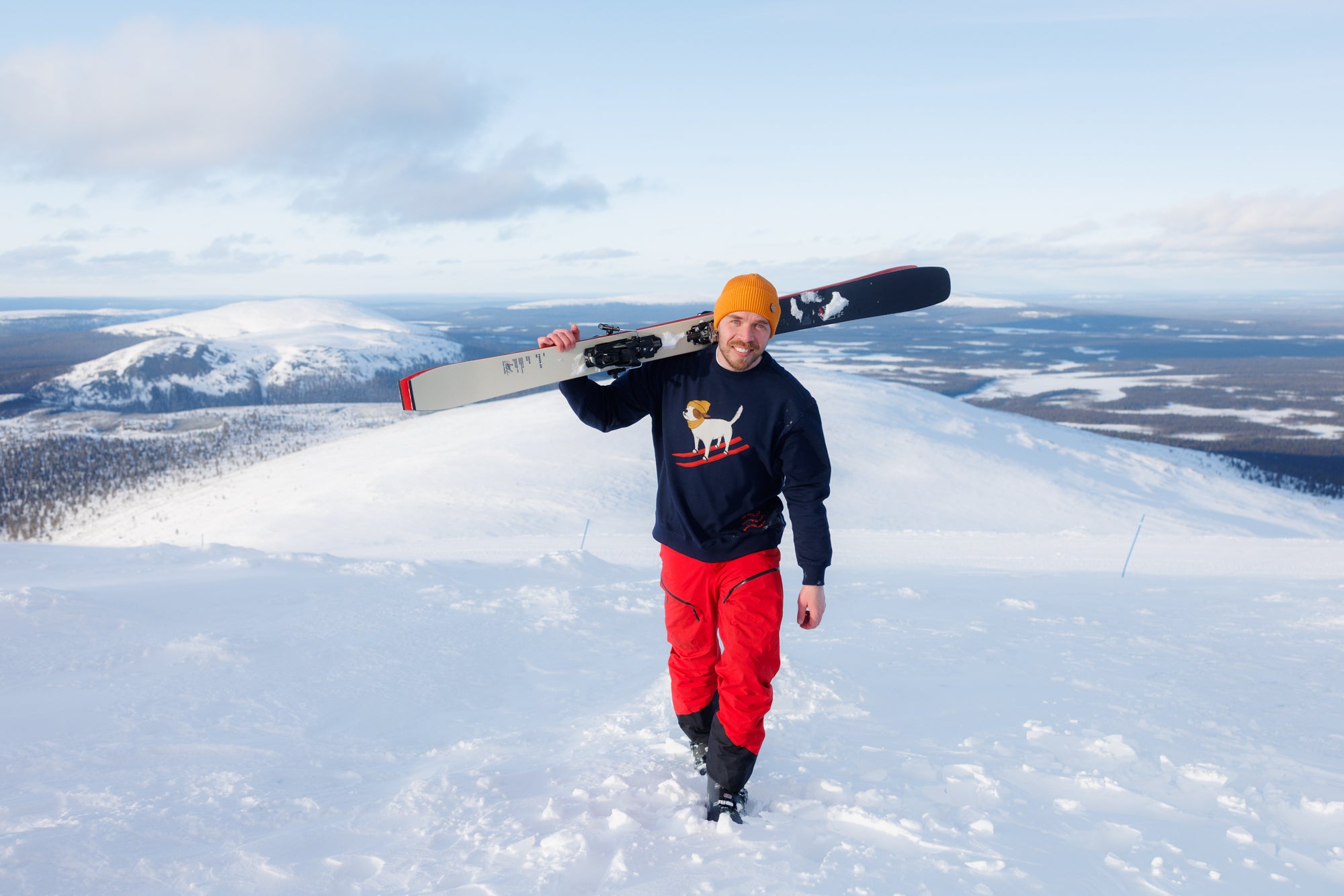 Man in red pants and yellow beanie carries skis on snowy mountain with scenic winter landscape in background.