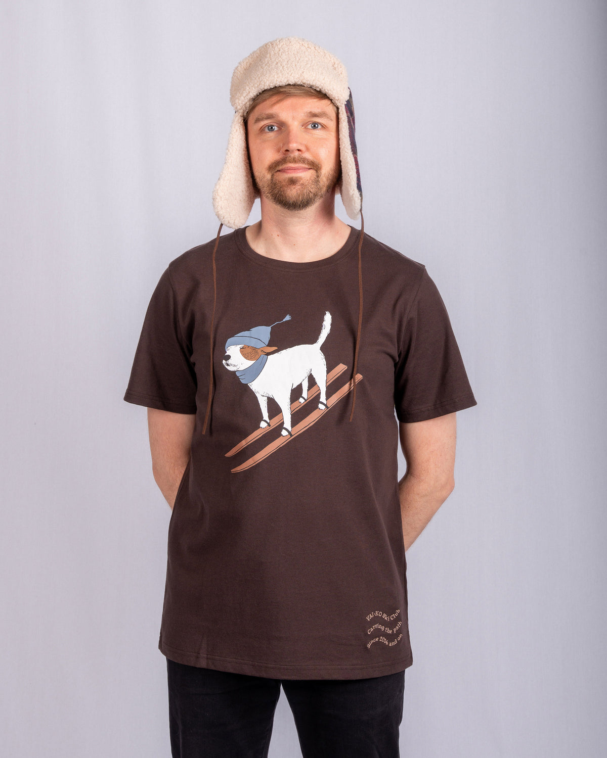 Man in a winter hat and brown t-shirt with a skiing dog graphic stands against a plain light background.
