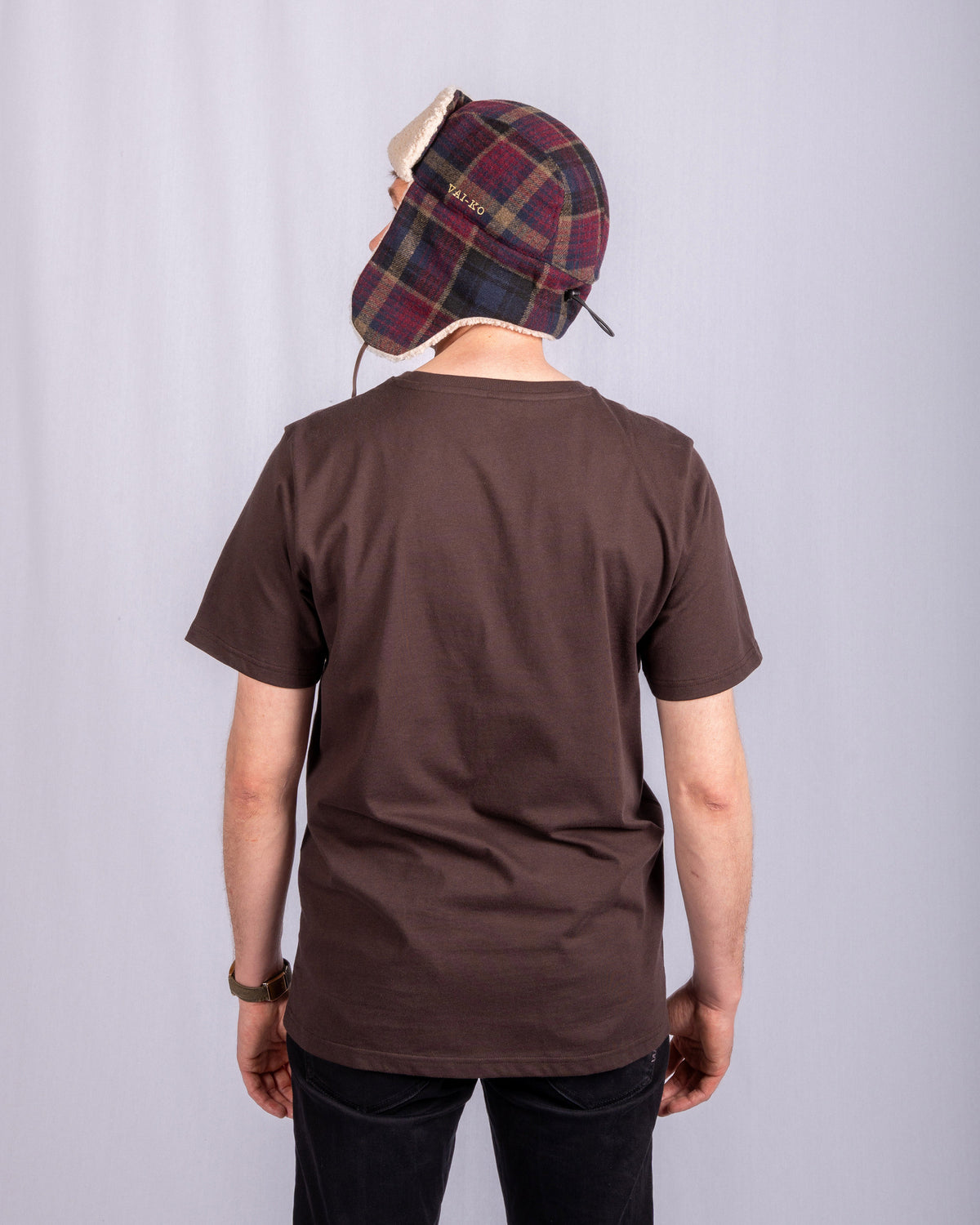 Person in a brown T-shirt and plaid trapper hat stands facing away against a plain light background.