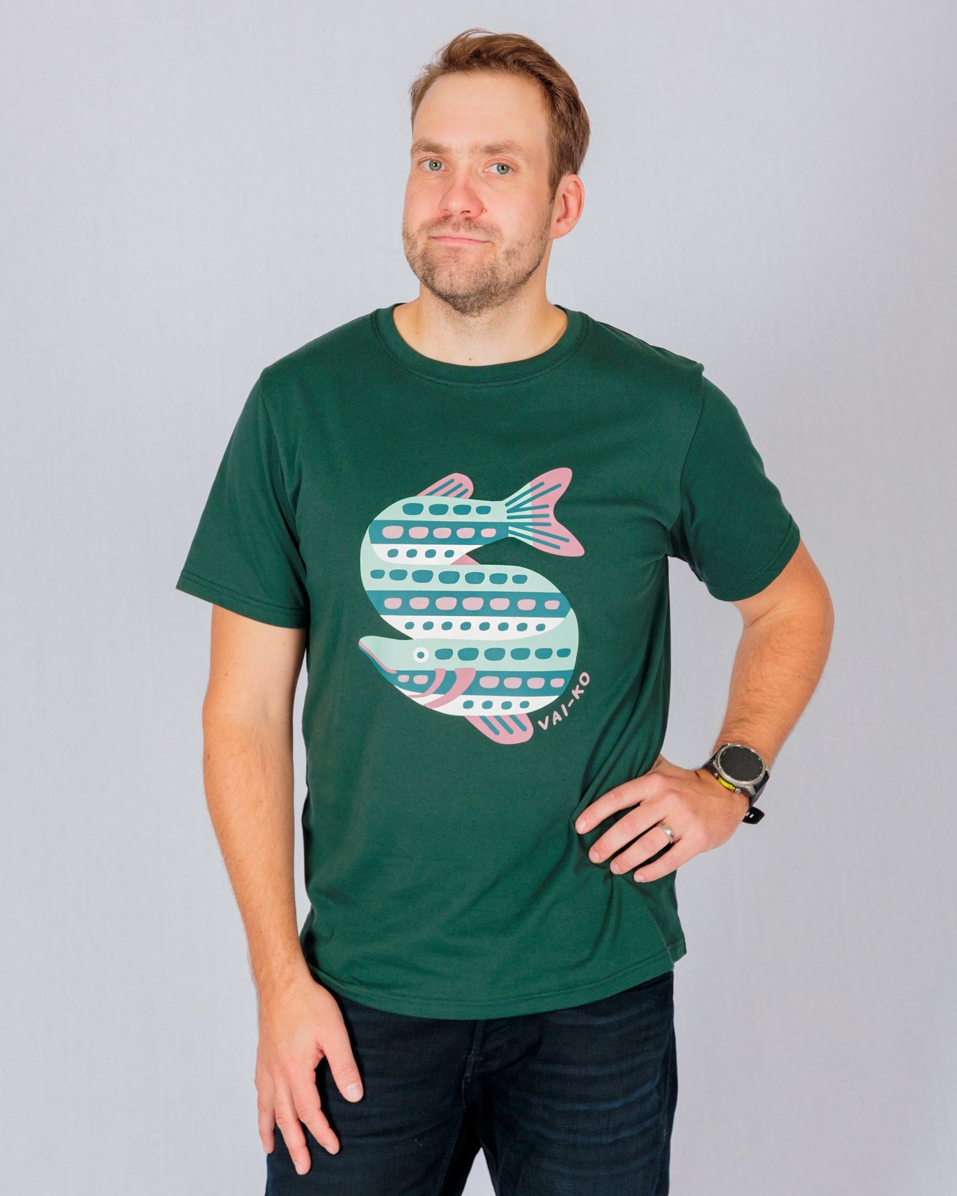 Man in a dark green t-shirt with a fish graphic, posing with one hand on his hip against a plain background.