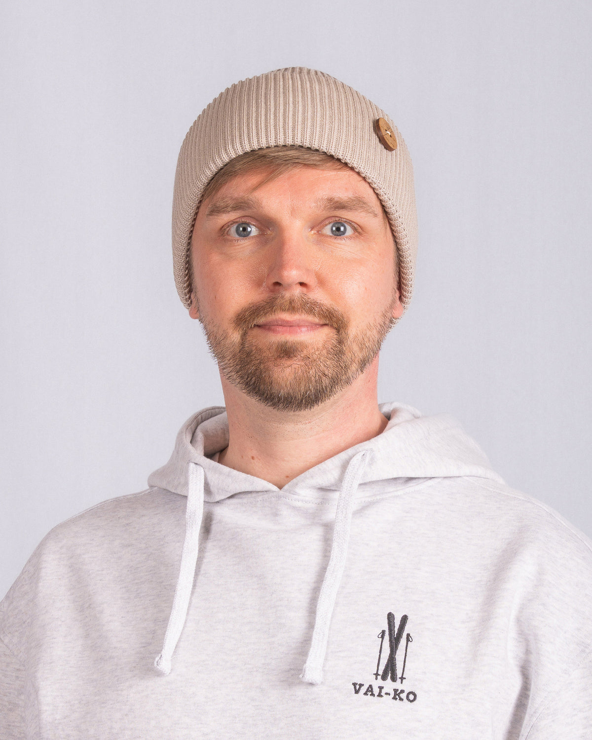 Man in beige knit beanie and light hoodie with VAI-KO logo, facing camera against plain background.